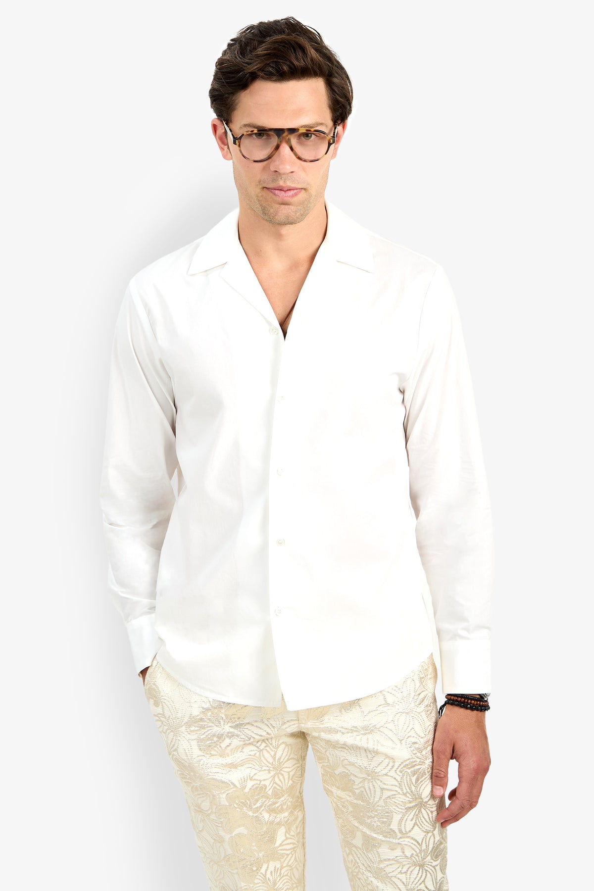 A man wearing glasses and the Able Shirt—a white, long-sleeve button-up—paired with light leaf-patterned pants stands against a plain white background with his hands in his pockets.