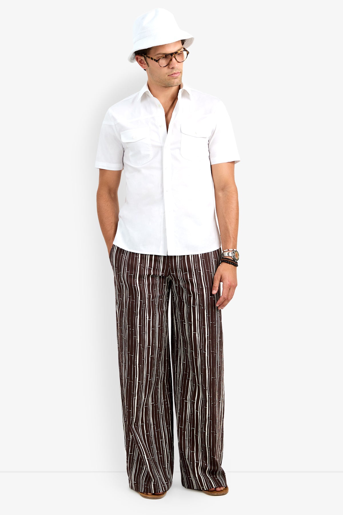 A man stands facing forward with his hands in his pockets, wearing the Shelton Short Sleeve Shirt in white, a white hat, glasses, and brown wide-leg pants with vertical white stripes.