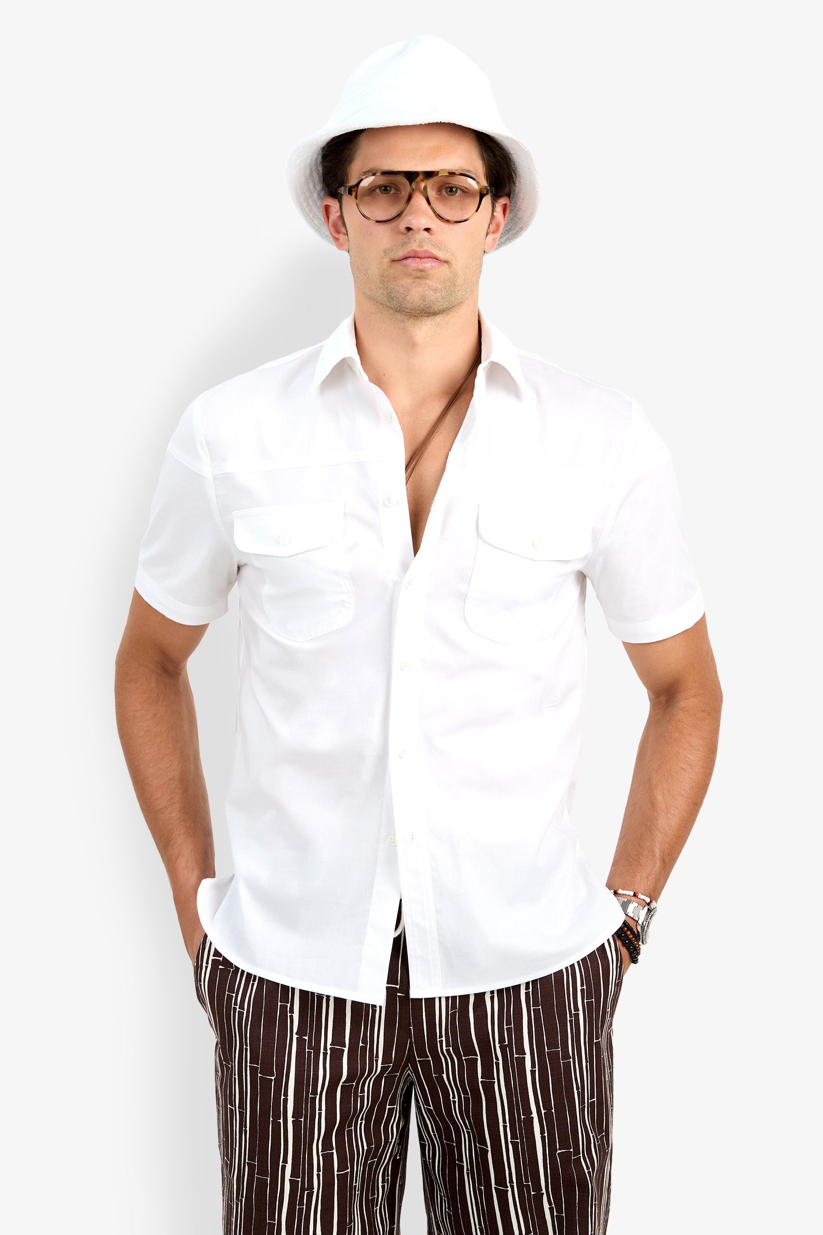 A man stands with his hands in his pockets, wearing glasses, a white hat, patterned brown and white pants, and the Shelton Short Sleeve Shirt against a plain light background.