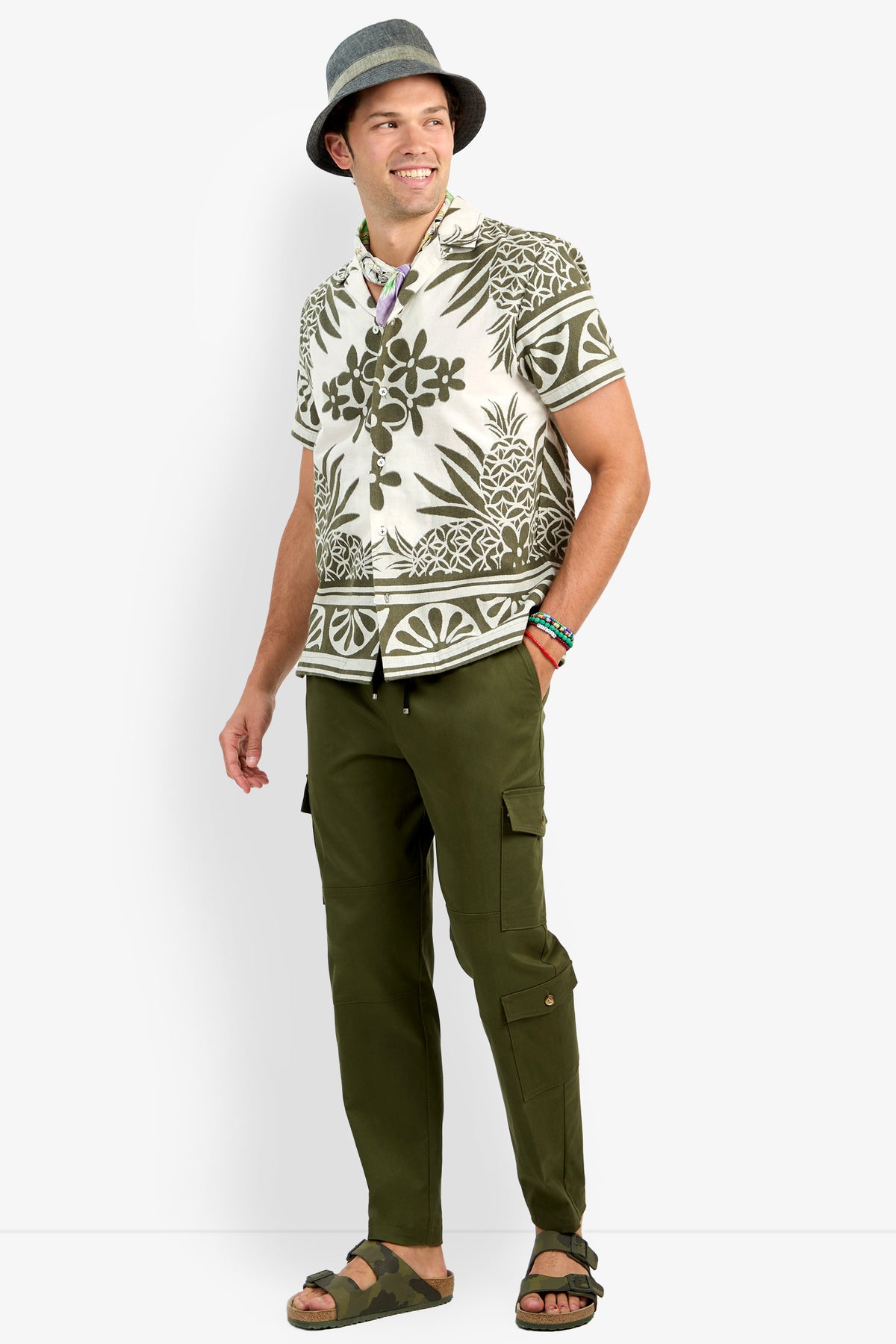 A smiling man wears the Felix Shirt, olive cargo pants, sandals, a gray hat, colorful bracelets, and a scarf around his neck as he stands casually against a plain background.