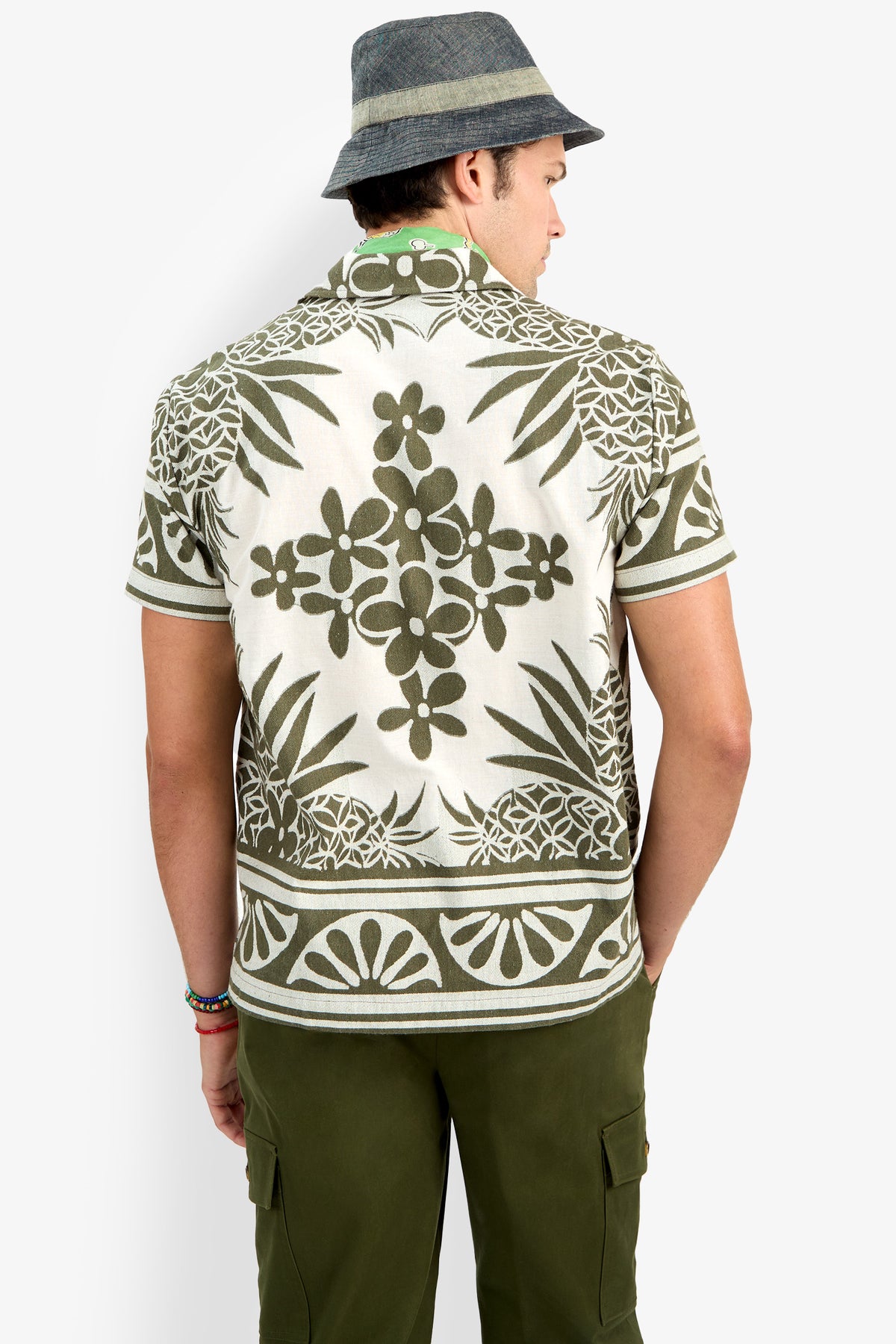 A person seen from behind wears a gray bucket hat, olive green cargo pants, and the Felix Shirt, which features a short-sleeve design with an olive green and white floral pattern.