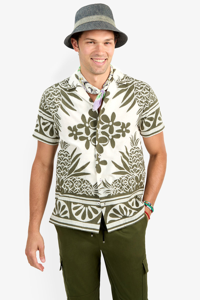 A man wearing the Felix Shirt, featuring green and white tropical patterns, along with a gray hat, olive green pants, a colorful neck scarf, and bracelets, smiles in front of a plain white background.