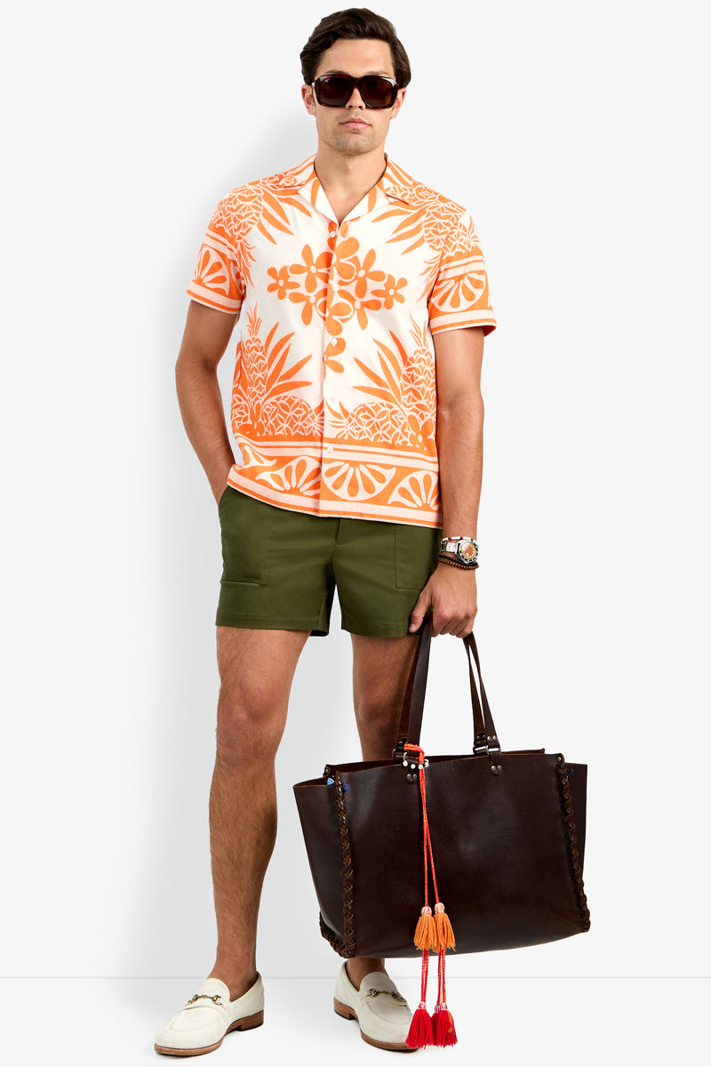 A man wearing sunglasses, the Felix Shirt, olive green shorts, white loafers, and holding a large dark brown tote bag with red and orange tassels stands confidently.