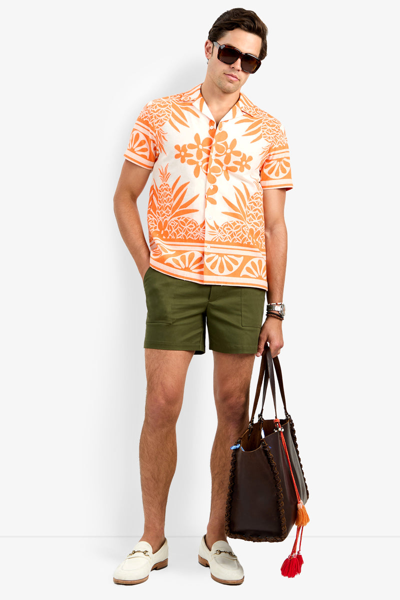 A man wearing sunglasses, the Felix Shirt (an orange and white patterned short-sleeve), olive green shorts, white loafers, and holding a brown tote bag with tassels stands against a plain white background.