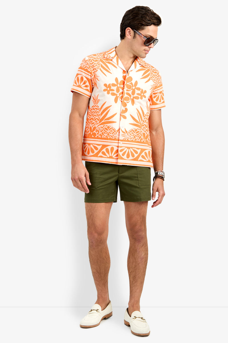 A man wearing sunglasses, an orange and white patterned short-sleeve shirt, olive green Will Rogers Shorts, and white loafers stands against a plain white background.