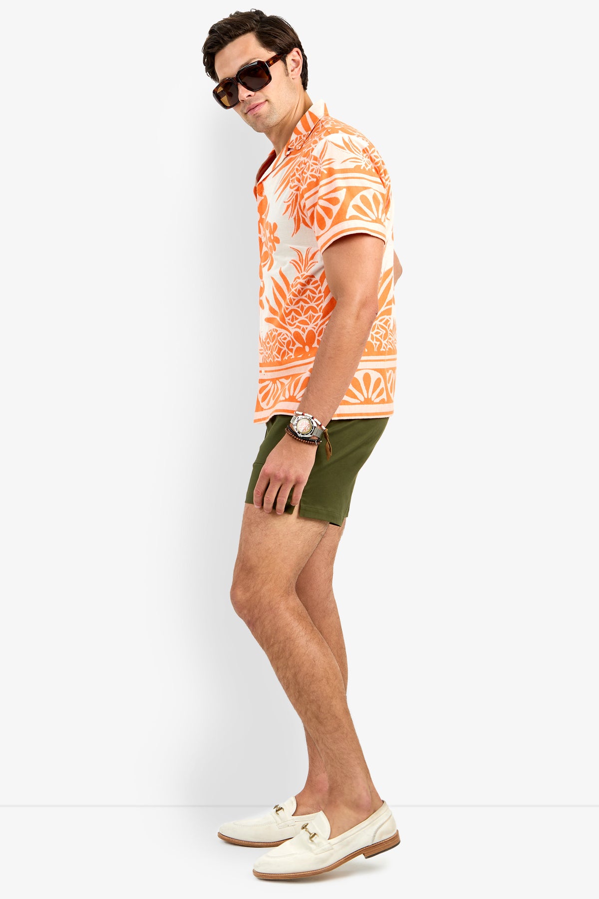 A man poses sideways in the Felix Shirt, featuring an orange floral print, paired with olive green shorts, white loafers, sunglasses, and a watch against a plain white background.