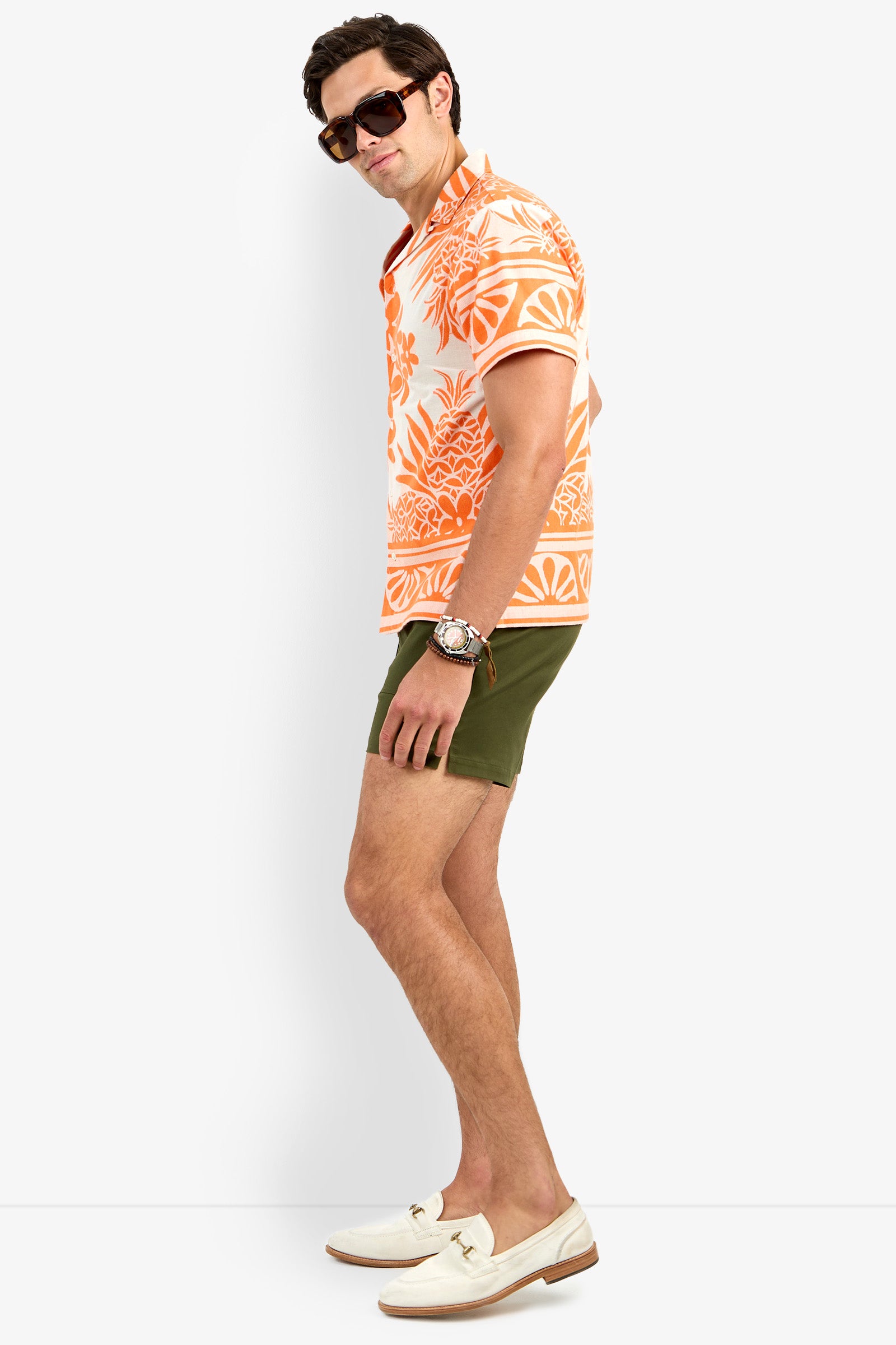 A man poses sideways in the Felix Shirt, featuring an orange floral print, paired with olive green shorts, white loafers, sunglasses, and a watch against a plain white background.