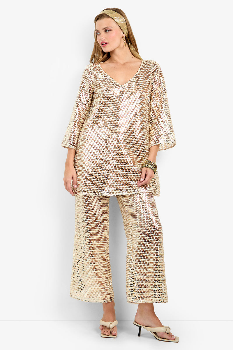 A woman models the Dariya Pant, a shiny gold sequin wide-leg pant paired with a loose V-neck top. She completes the look with gold sandals, a matching headband, bracelets, and her hair styled down to the side.