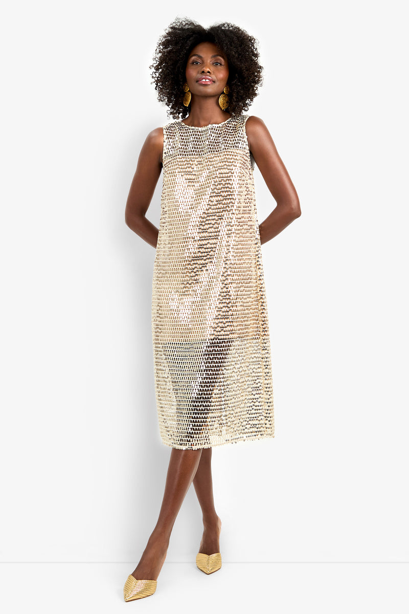 A woman poses against a white background in the Eleanor Dress, a sleeveless, knee-length gold sequin design. She pairs it with gold earrings, pointed-toe gold shoes, and voluminous curls.