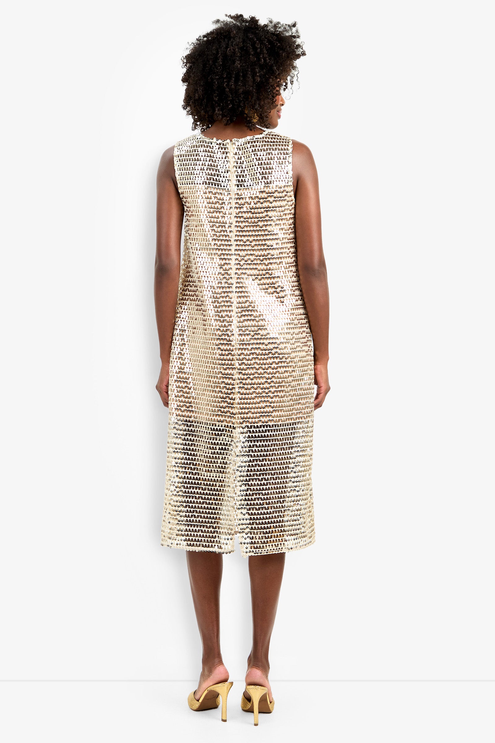 A person with curly hair, seen from behind, wears the sleeveless, knee-length gold sequin Eleanor Dress and light yellow heeled sandals against a plain white background.