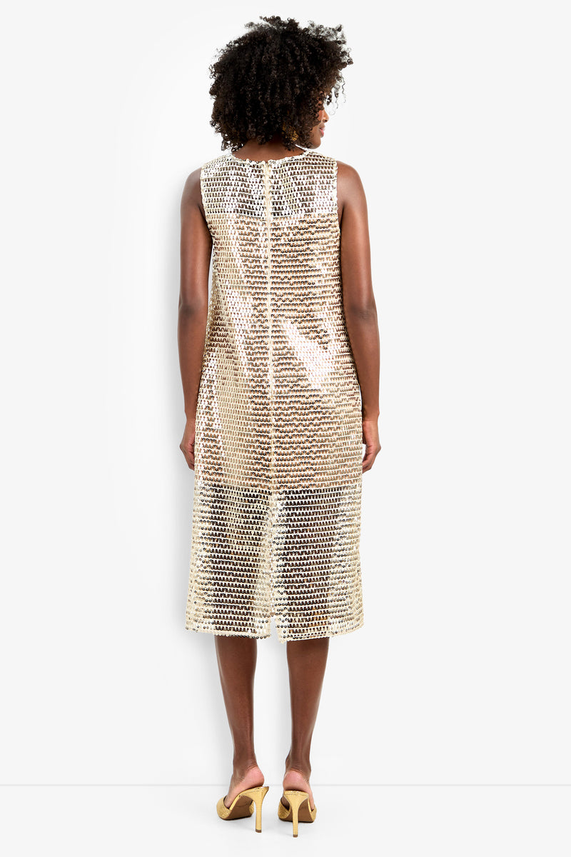 A person with curly hair, seen from behind, wears the sleeveless, knee-length gold sequin Eleanor Dress and light yellow heeled sandals against a plain white background.