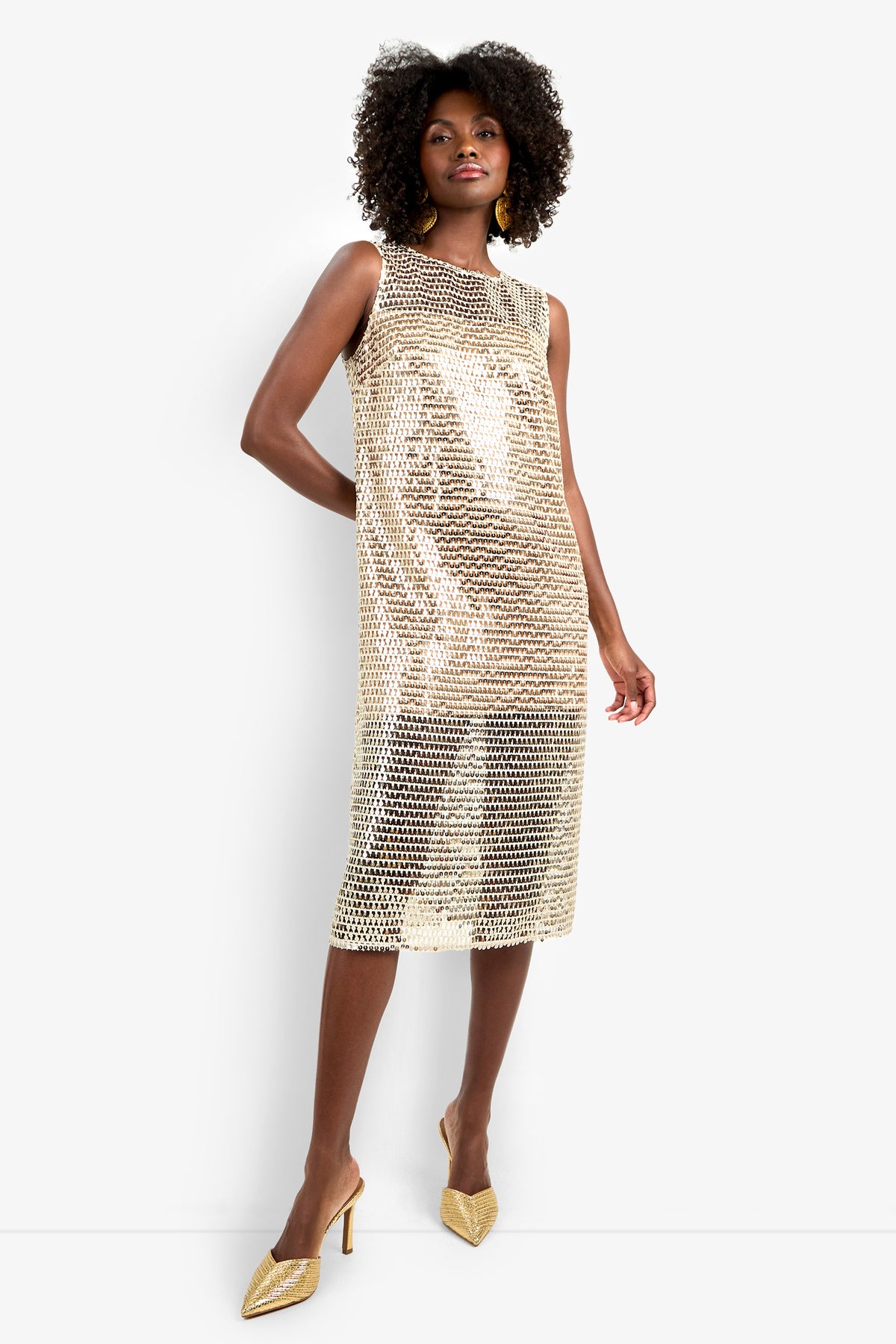 A woman stands confidently in the Eleanor Dress, a sleeveless, knee-length gold sequin mesh dress over a nude slip, paired with gold pointed-toe heels and large gold earrings. Her curly hair adds to the elegant look.