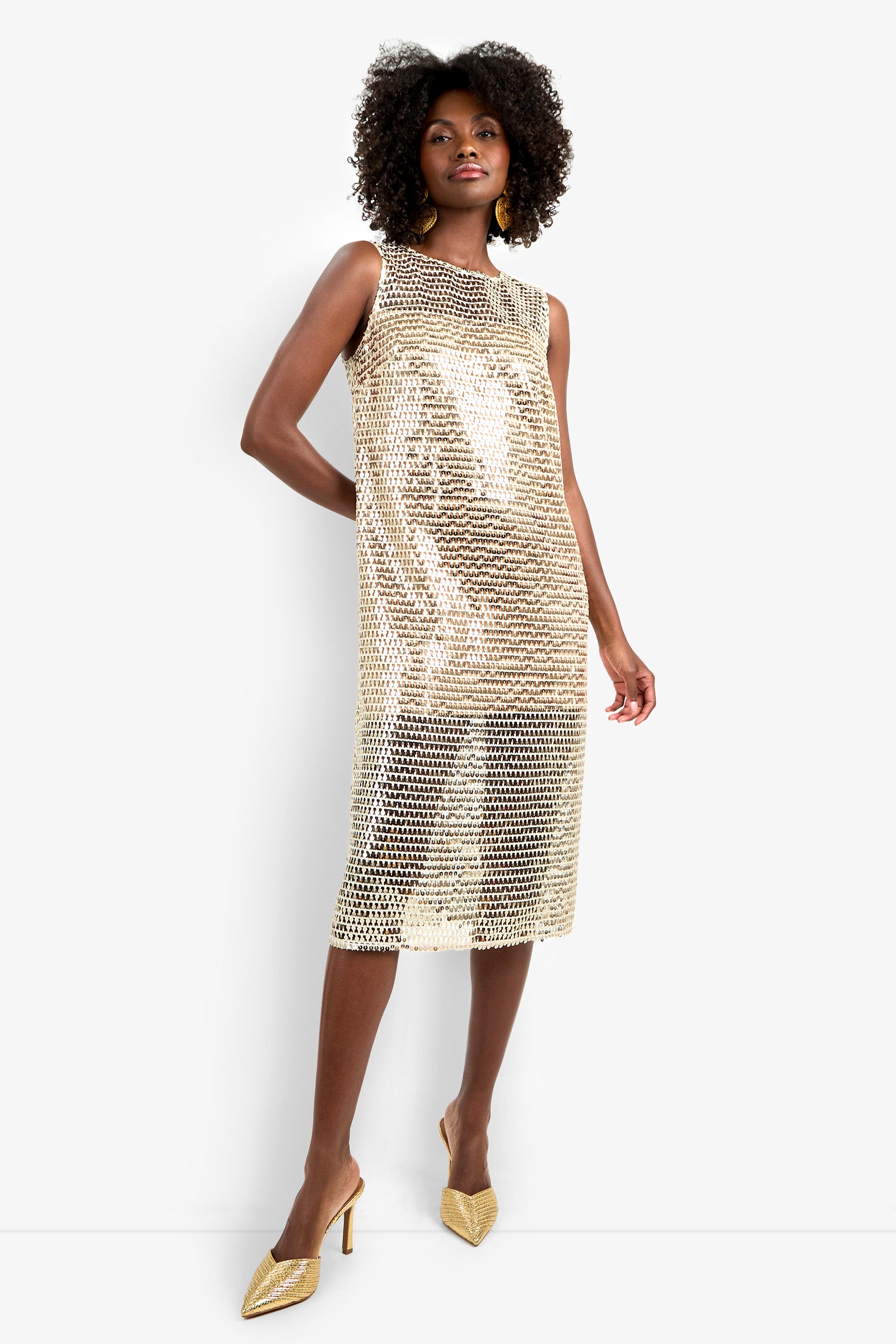 A woman stands confidently in the Eleanor Dress, a sleeveless, knee-length gold sequin mesh dress over a nude slip, paired with gold pointed-toe heels and large gold earrings. Her curly hair adds to the elegant look.