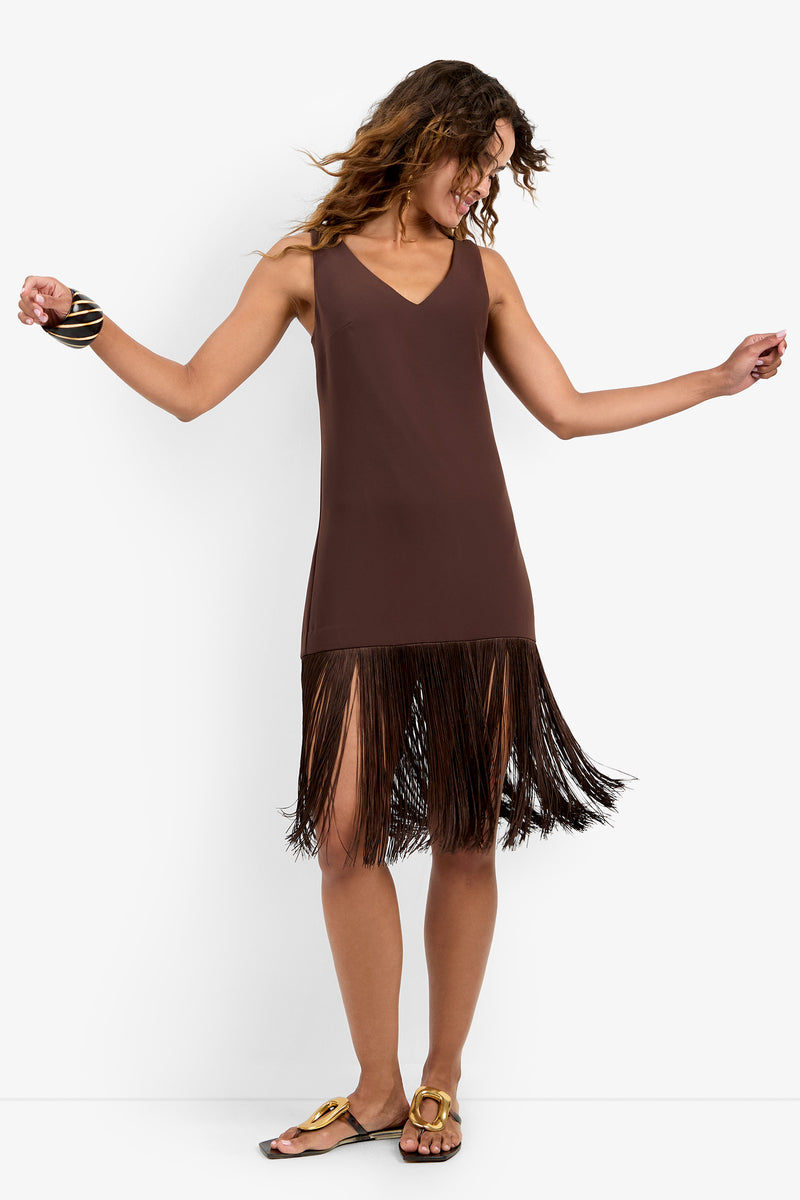 A woman in the AYMANI DRESS—a sleeveless brown polyester dress with fringe at the hem—dances, smiling with her head turned sideways. She has wavy hair, wears sandals, and a chunky bracelet on her left wrist.