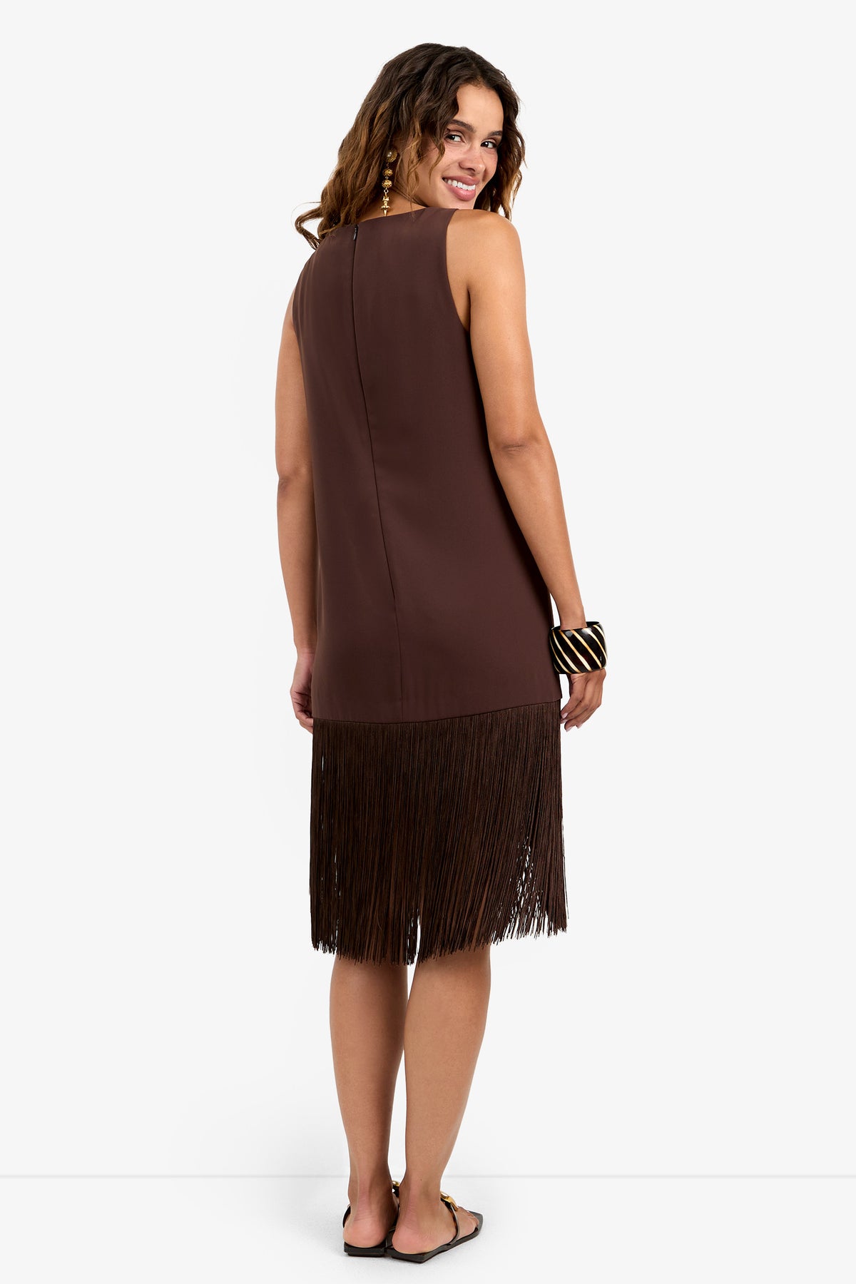 A woman stands against a plain white background, smiling over her shoulder and wearing the sleeveless brown Aymani Dress with long fringe at the hem, black sandals, and a chunky black and white bracelet.