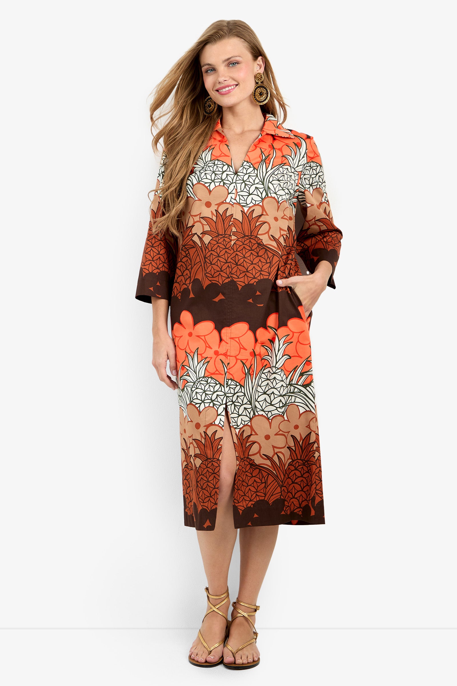 A woman stands smiling in the Kamia Dress, a knee-length piece featuring a bold orange, brown, and white floral-leaf print. The dress has a collar, three-quarter sleeves, handy pockets, and is paired with sandals and gold earrings.