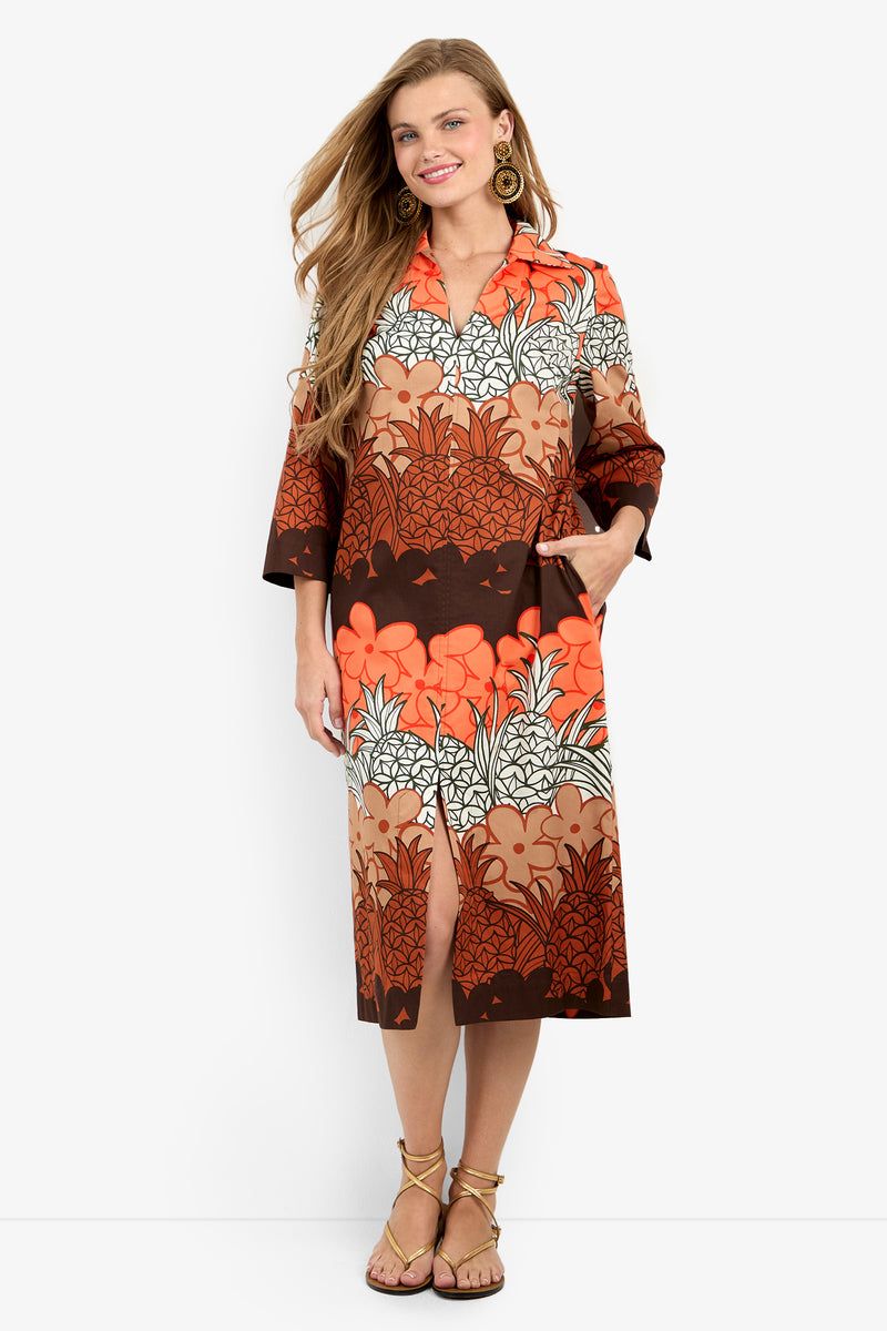 A woman stands smiling in the Kamia Dress, a knee-length piece featuring a bold orange, brown, and white floral-leaf print. The dress has a collar, three-quarter sleeves, handy pockets, and is paired with sandals and gold earrings.