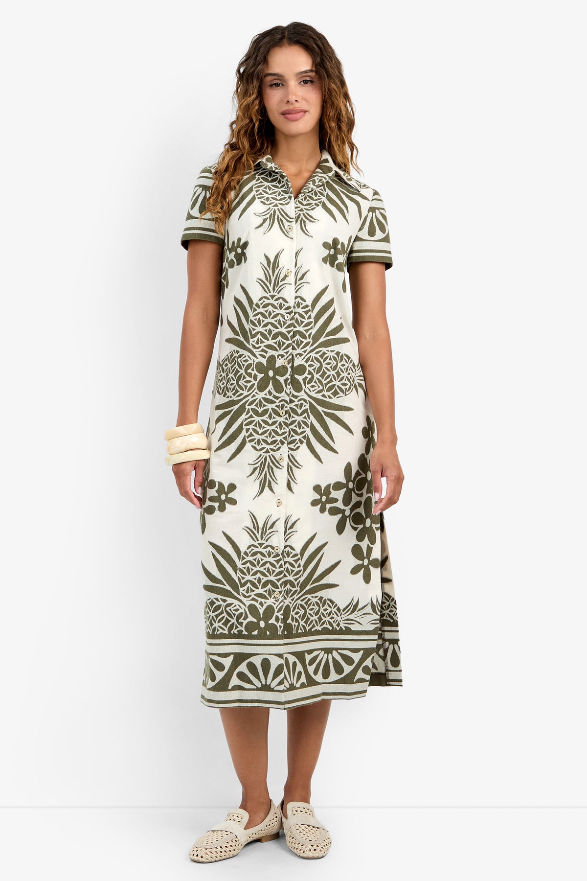 A woman in the Abba Dress—a cream, short-sleeve collared midi with green tropical pineapple and leaf prints—paired with beige flat woven shoes and chunky bangles, stands against a white background.