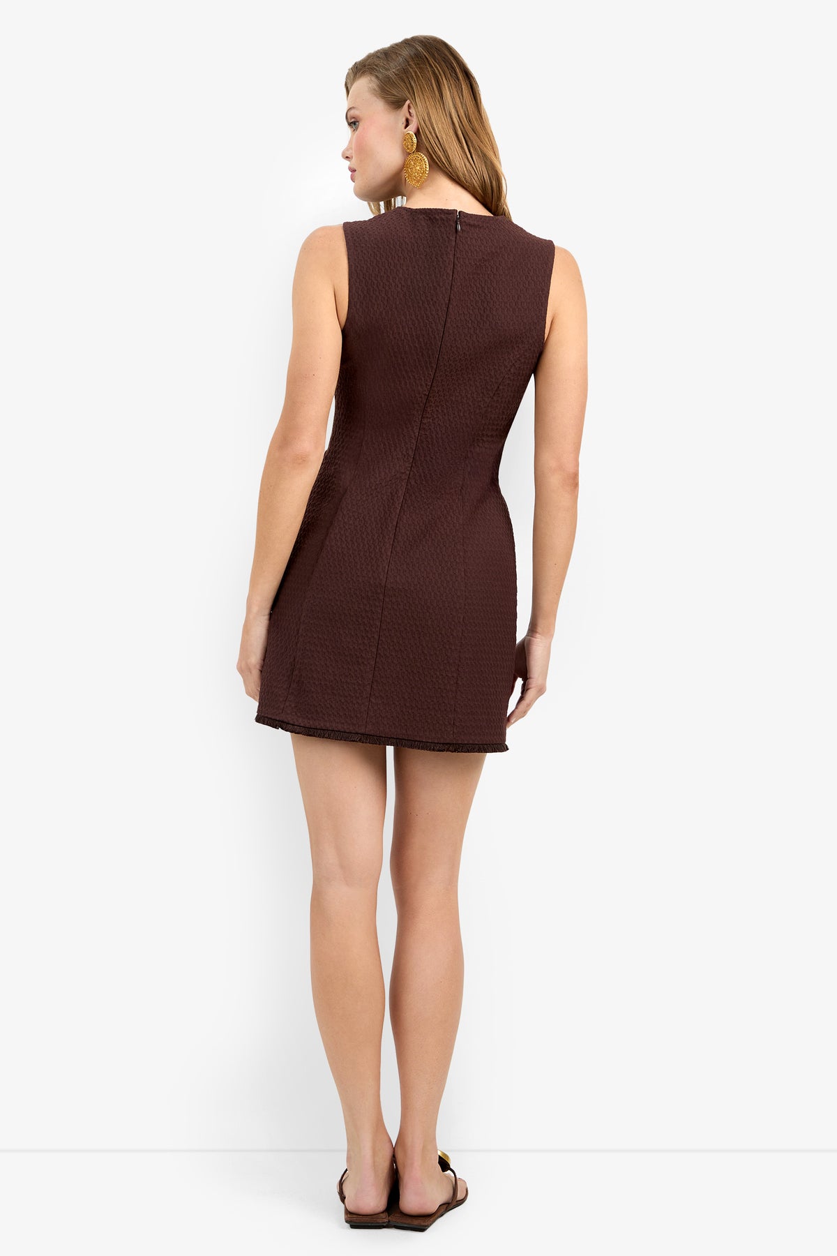 A woman stands with her back to the camera, wearing the Dame Fringe Dress—a sleeveless, textured brown mini dress—paired with black sandals and gold earrings, against a plain white background.