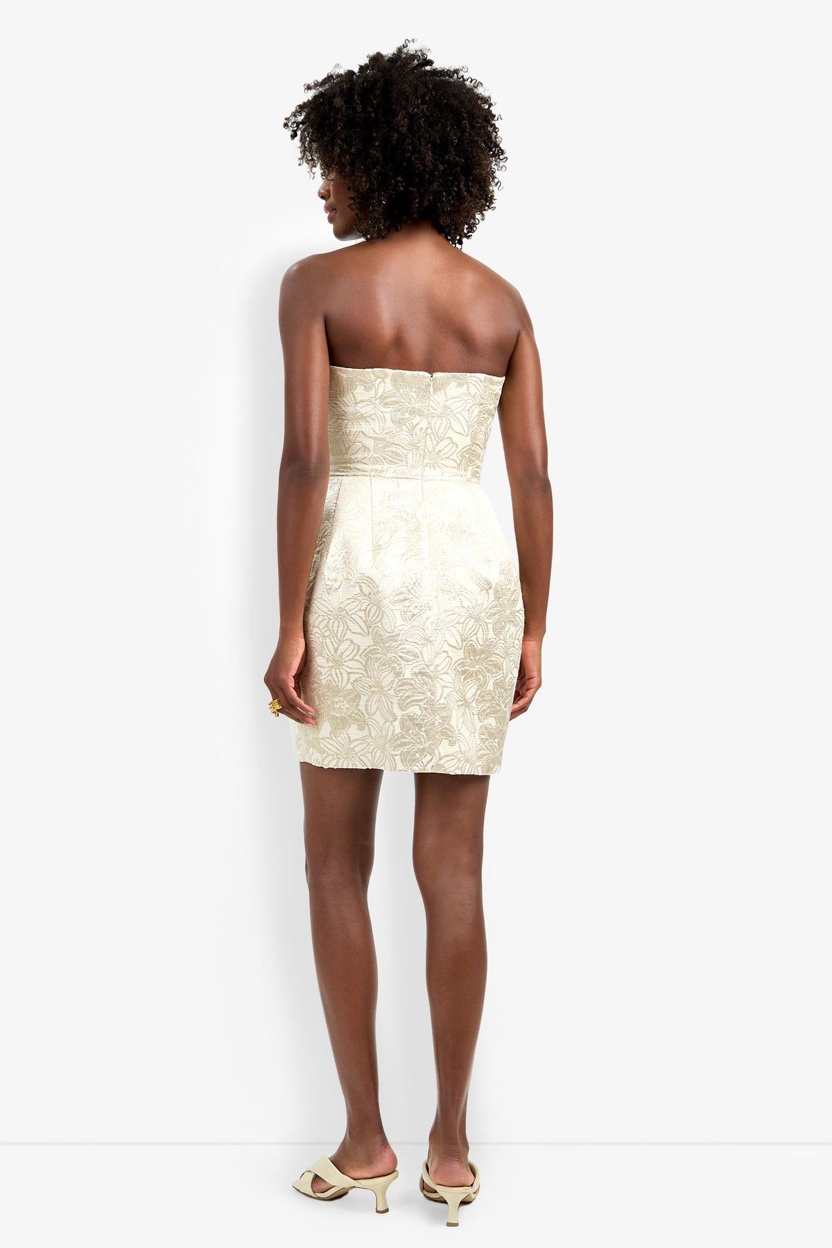 A woman with short curly hair stands with her back to the camera, wearing the strapless cream floral Kalley Dress paired with matching open-toe heels.