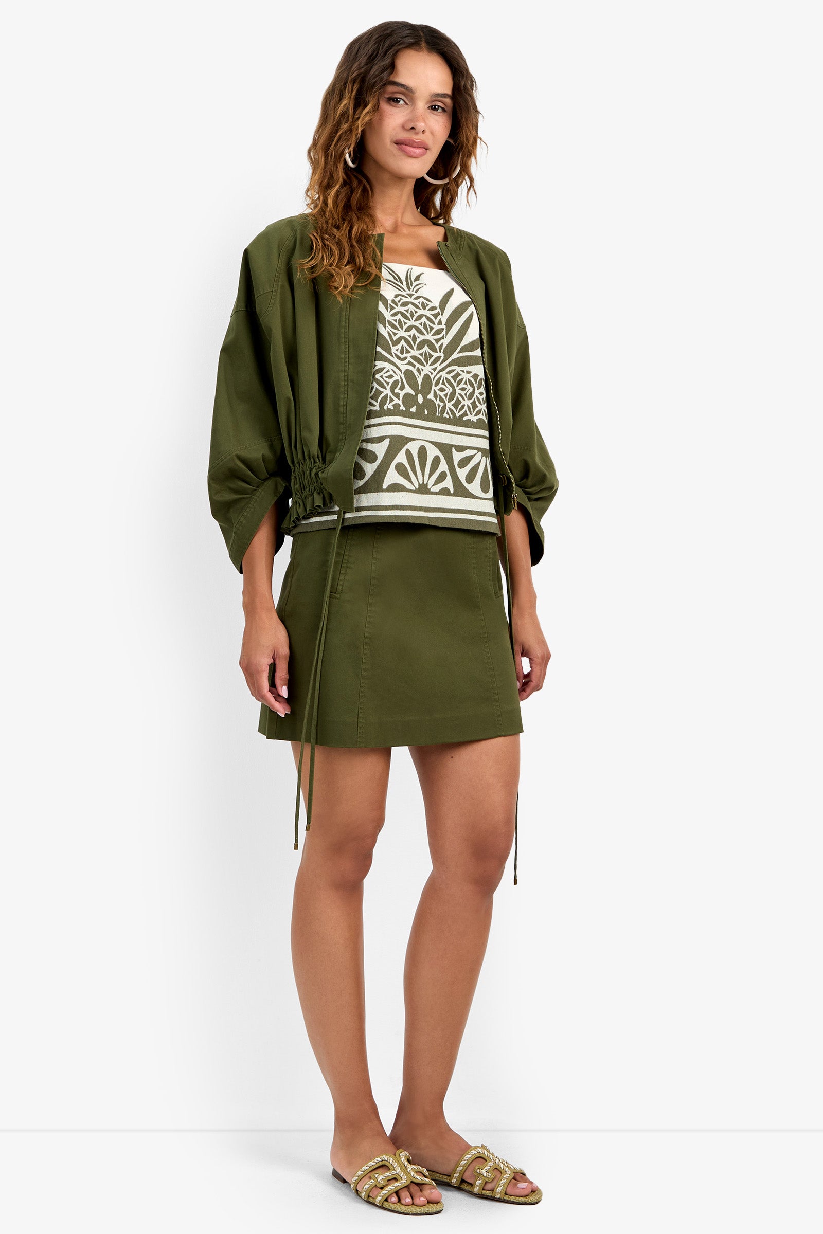 A woman with wavy hair wears the Prita Jacket in olive green over a patterned white and green top and matching skirt, paired with slide sandals. The background is plain white.