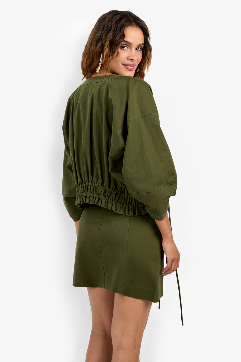 A woman with wavy hair smiles over her shoulder, wearing the Prita Jacket—an olive green, long-sleeve top with a gathered waistband—paired with a matching mini skirt against a plain light background.