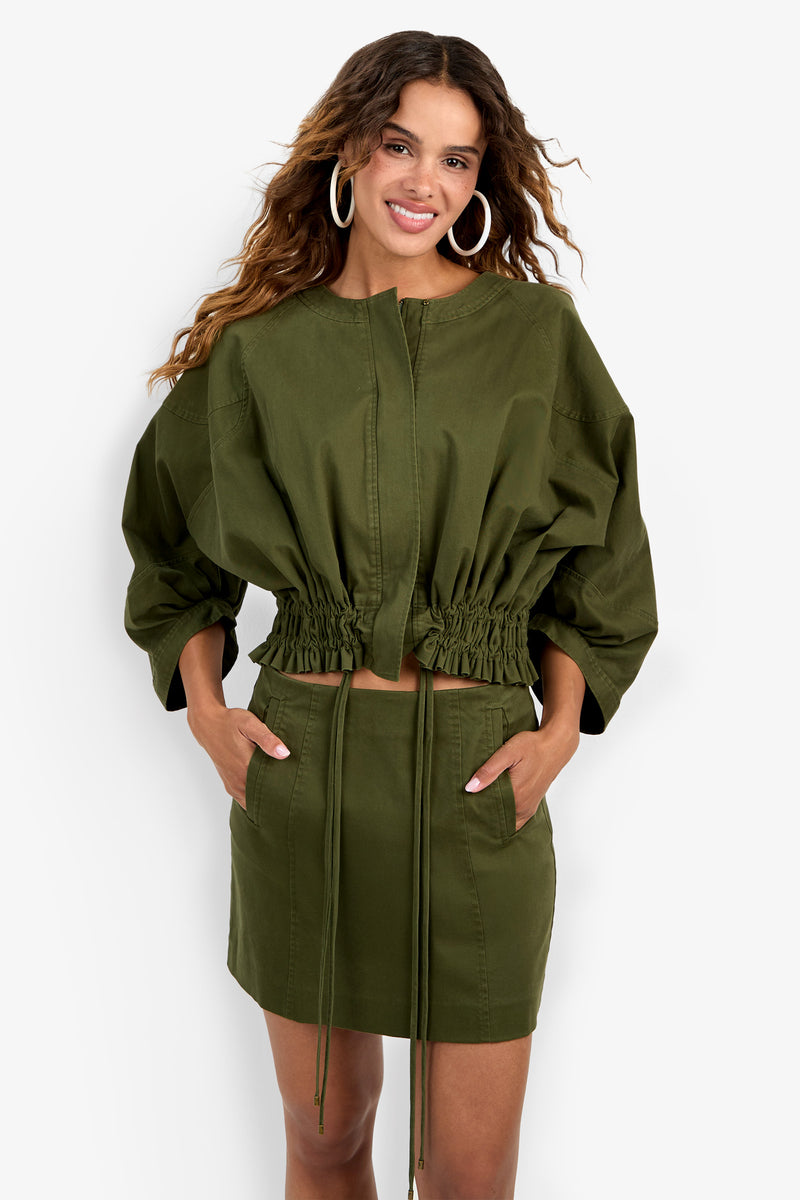 A woman wearing the Prita Jacket in olive green, styled with a matching high-waisted mini skirt. She has long wavy hair, large hoop earrings, and her hands in her skirt pockets.