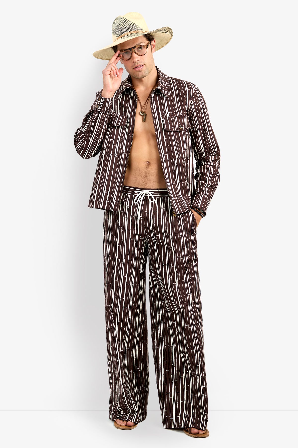 A man stands barefoot against a plain white background, wearing the Arturo Jacket—an open brown and white vertical striped jacket with matching wide-leg pants, accessorized with glasses and a wide-brimmed straw hat.