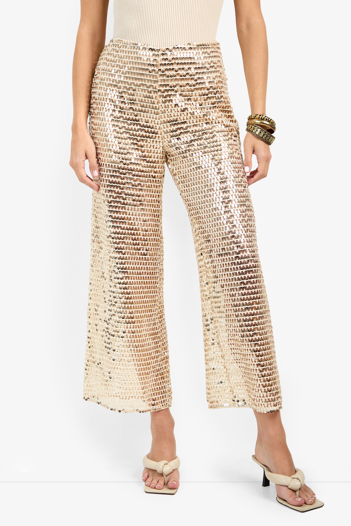 A woman wears the Dariya Pant—shiny, gold sequin wide-leg trousers—paired with a light ribbed top, beige heeled sandals with bows, and gold bangles on her wrist against a plain white background.