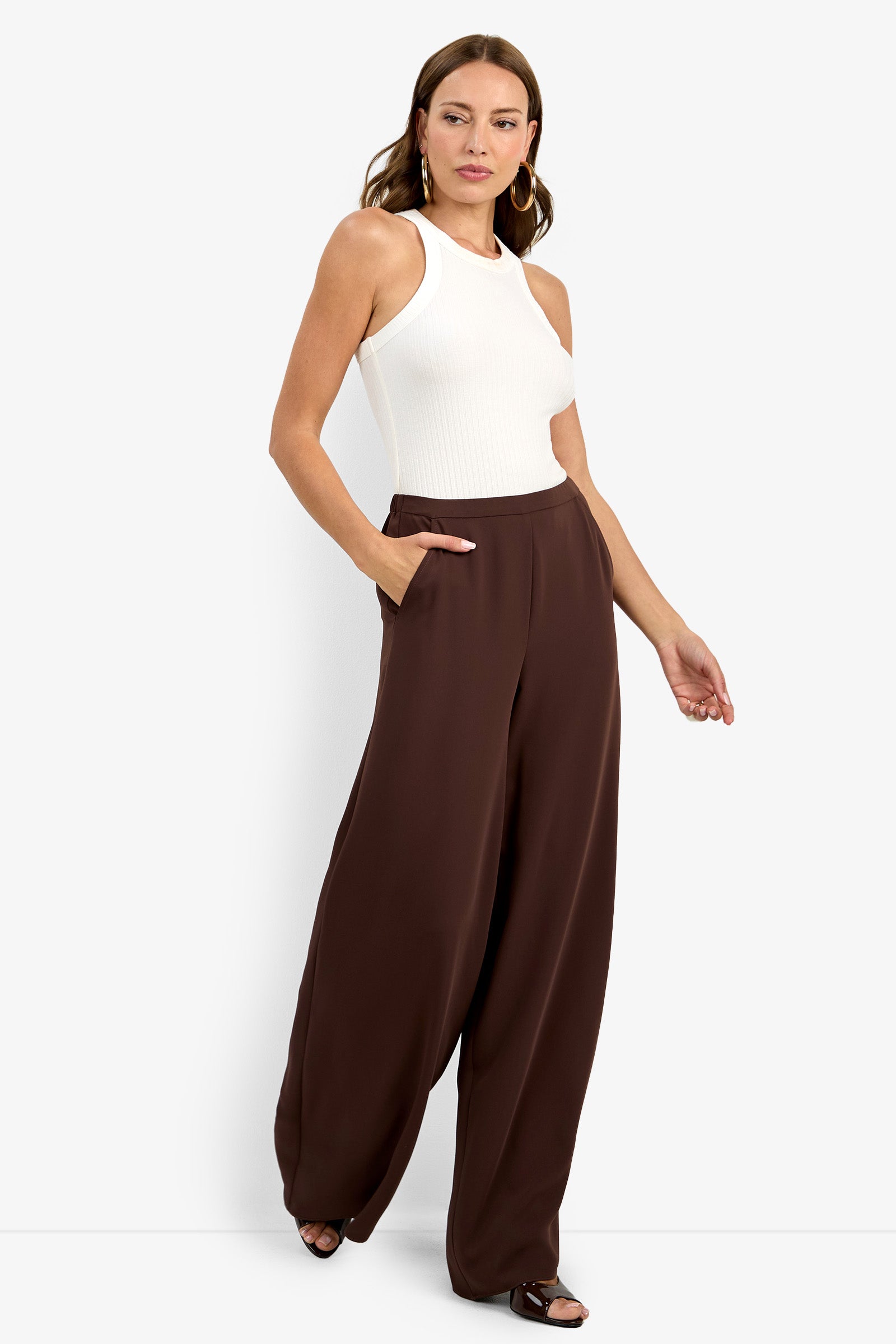 A woman stands against a white background wearing a sleeveless white ribbed top and Aloness Pant—high-waisted, wide-leg brown pants with pockets. She has loose hair, hoop earrings, and strappy black sandals.