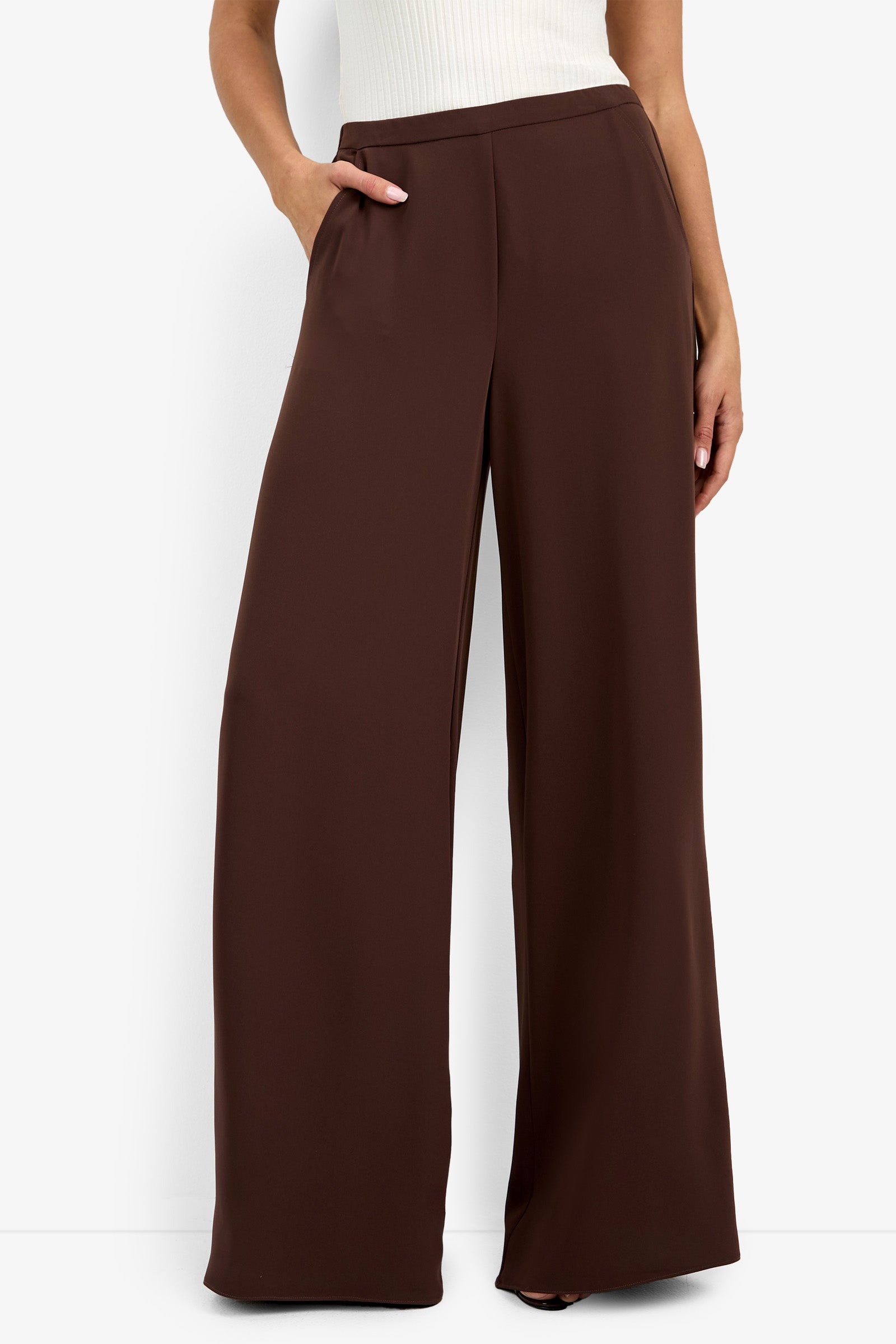 A person wearing the Aloness Pant, a pair of high-waisted, wide-leg brown trousers, and a sleeveless white top stands against a plain white background with one hand in their pocket.