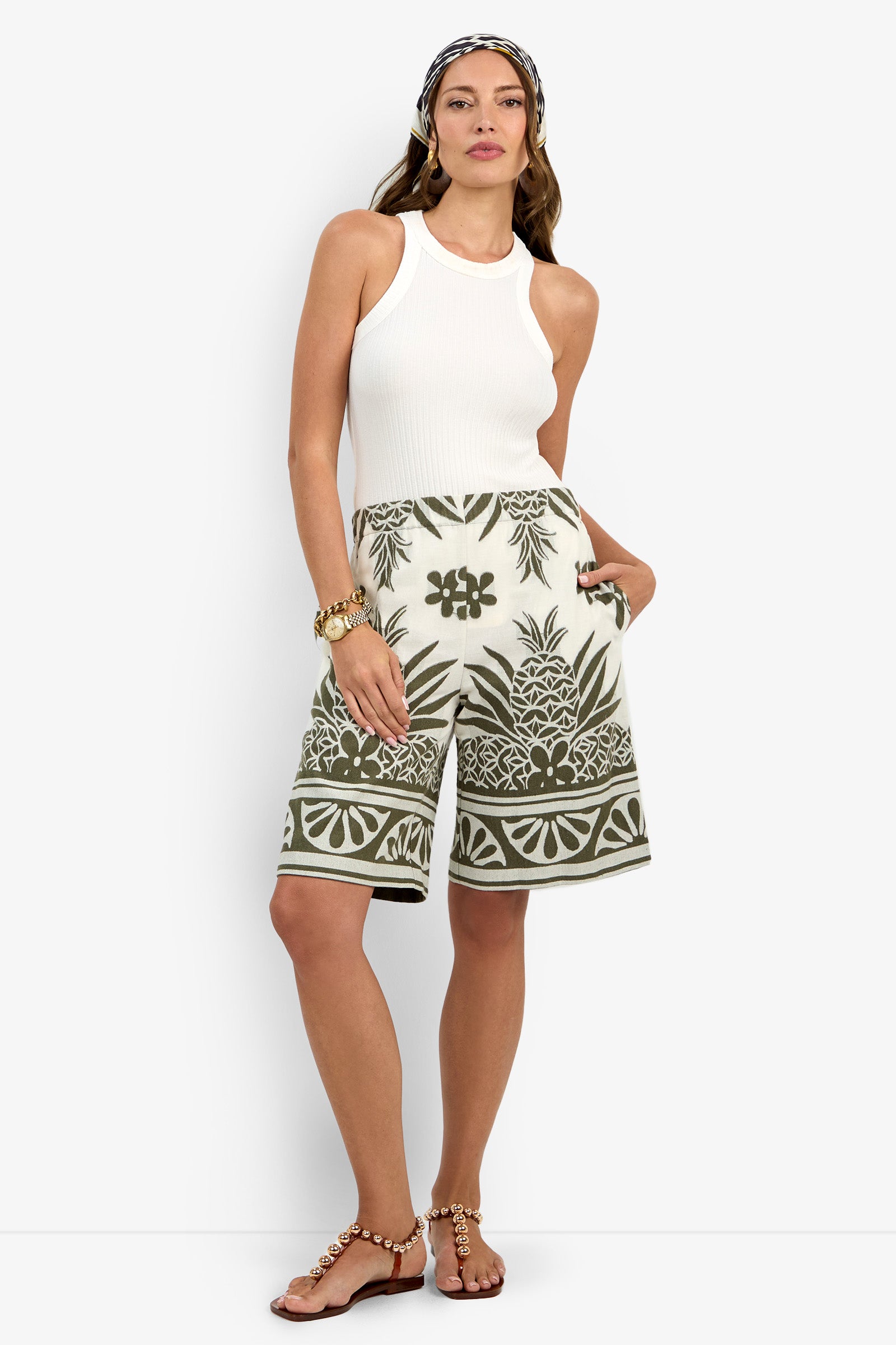 A woman wearing a white sleeveless ribbed tank top, the Estella Short featuring green tropical patterns, pearl-detailed sandals, gold jewelry, and a patterned headband stands against a white background.