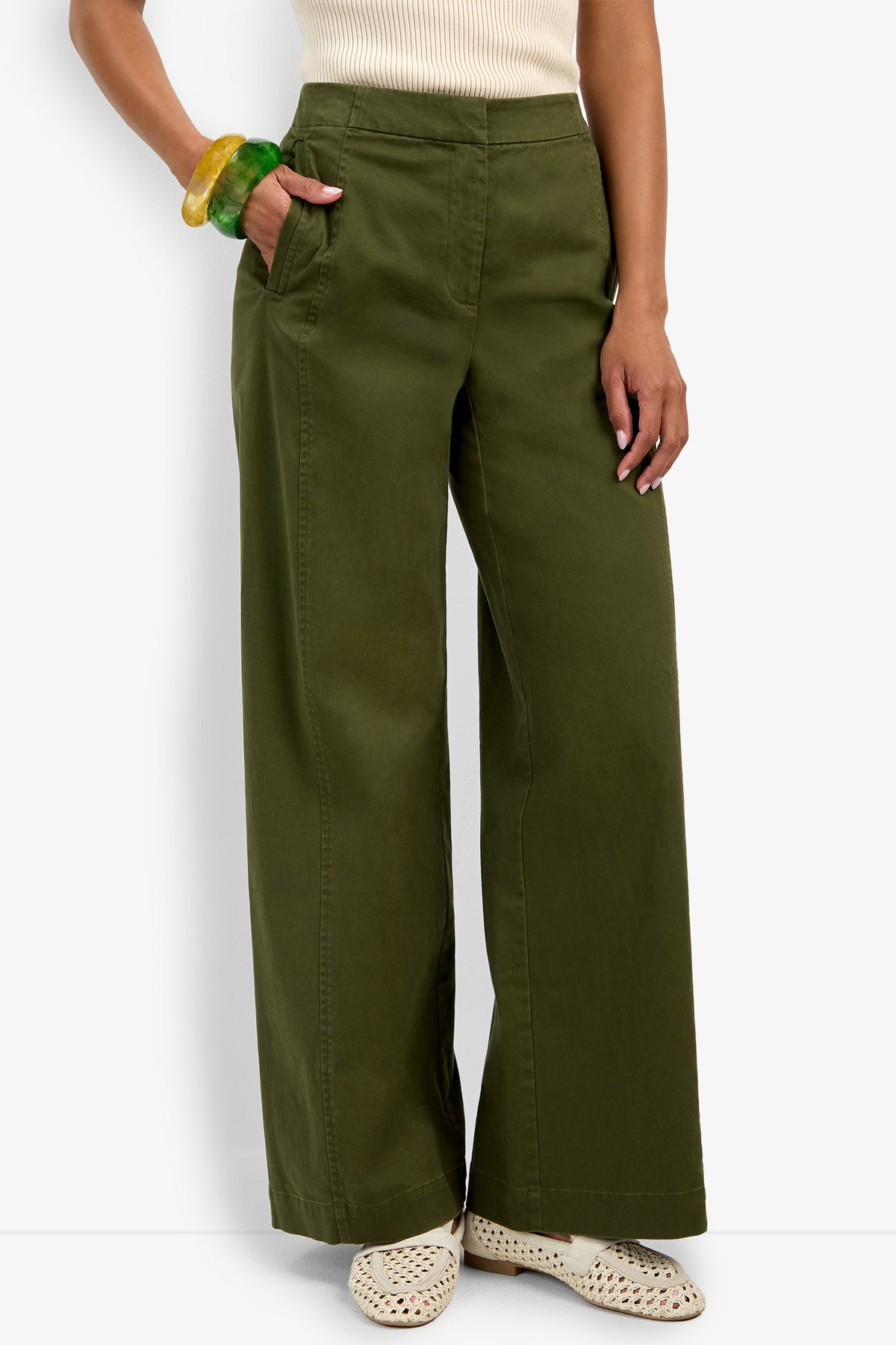 Wearing the Carum Pant—high-waisted, wide-leg olive green trousers—paired with a sleeveless cream top, woven beige shoes, and chunky green and yellow bangles, this person stands with one hand in their pocket.
