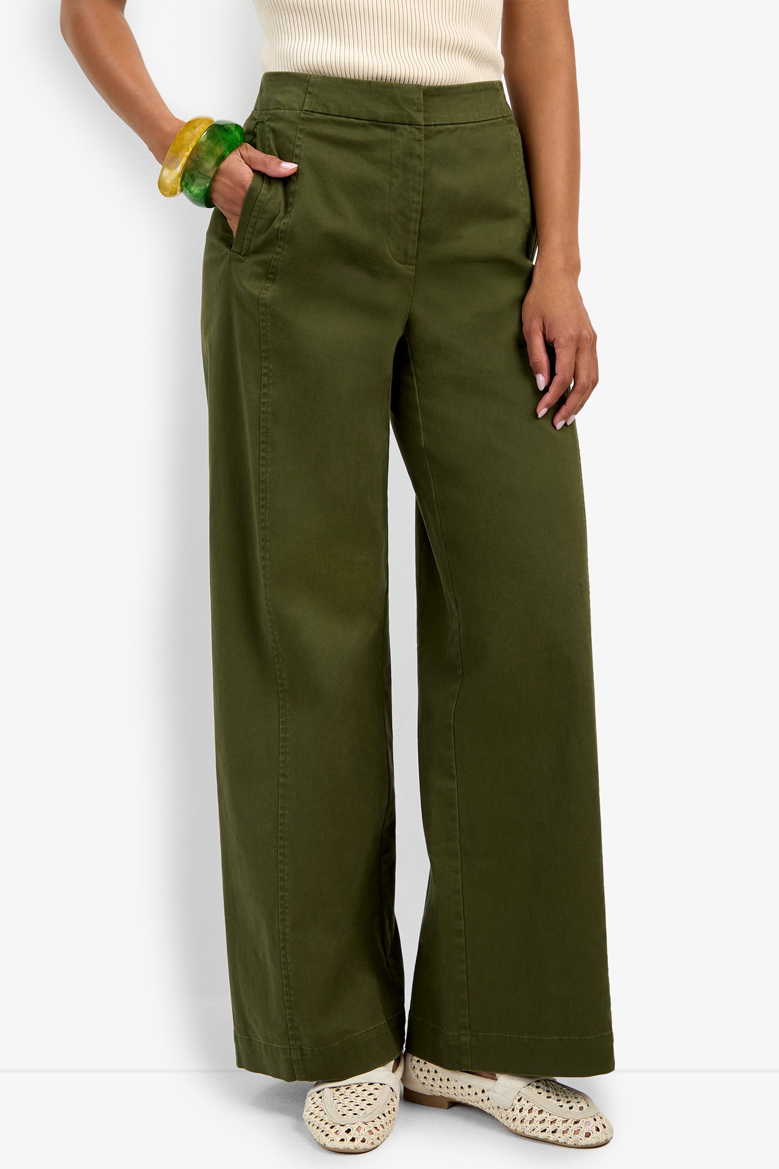 Wearing the Carum Pant—high-waisted, wide-leg olive green trousers—paired with a sleeveless cream top, woven beige shoes, and chunky green and yellow bangles, this person stands with one hand in their pocket.