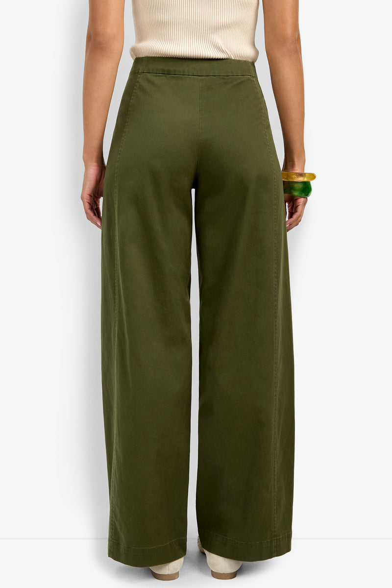 A person wearing the Carum Pant—high-waisted, wide-leg olive green pants—and a sleeveless beige top stands with their back to the camera, accessorized with two chunky bracelets on their right wrist.