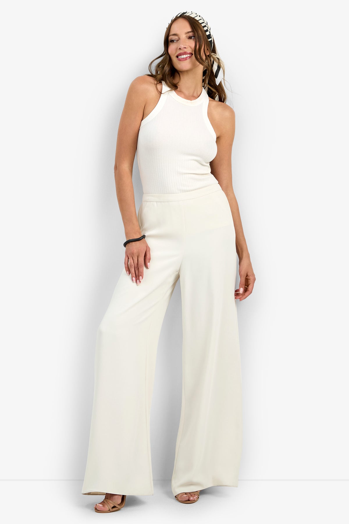 A woman smiles in the Aloness Pant—wide-leg, white pants—paired with a sleeveless white ribbed top, nude heels, long wavy brown hair, a black-and-white headband, and a black bracelet on her right wrist.