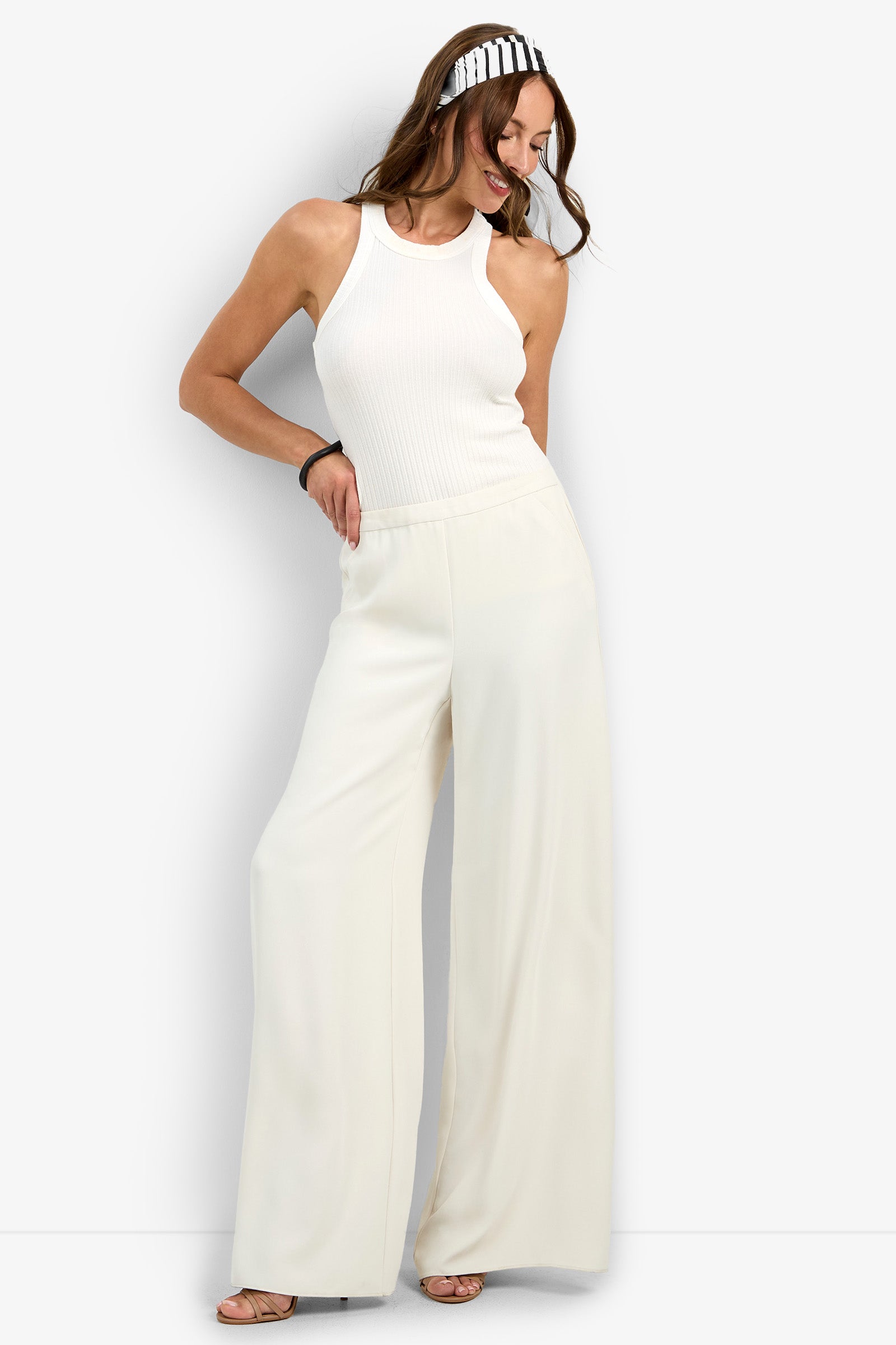 A woman stands smiling with one hand on her hip, wearing the Aloness Pant paired with a white sleeveless ribbed top, a black and white striped headband, and a black bracelet.
