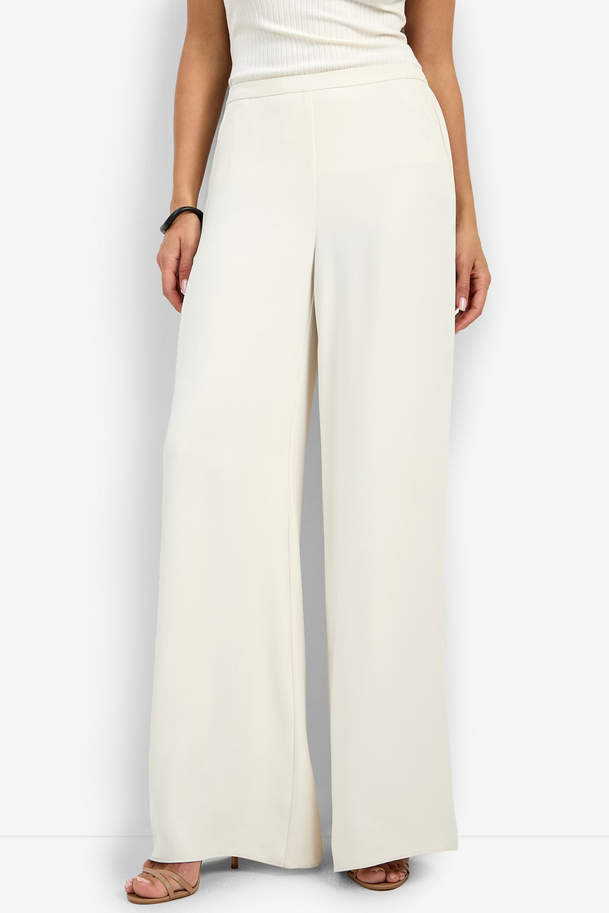 A person is shown from the shoulders down wearing the Aloness Pant—high-waisted, wide-leg cream-colored trousers—paired with a sleeveless white top and open-toe heeled sandals.