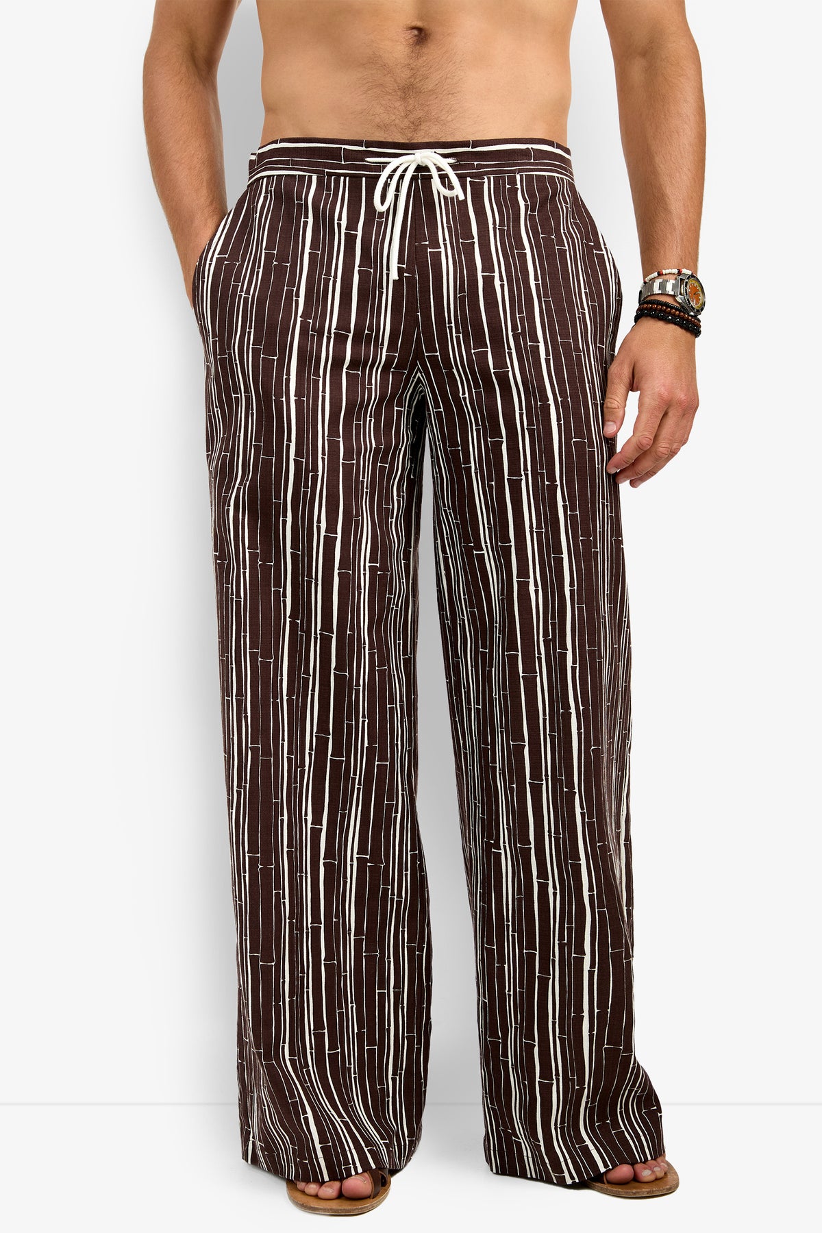 A man models the Wakiki Trouser—loose brown pants with vertical white stripes and a white drawstring waist—paired with sandals, shirtless, one hand in his pocket, bracelets and a watch on the other arm.