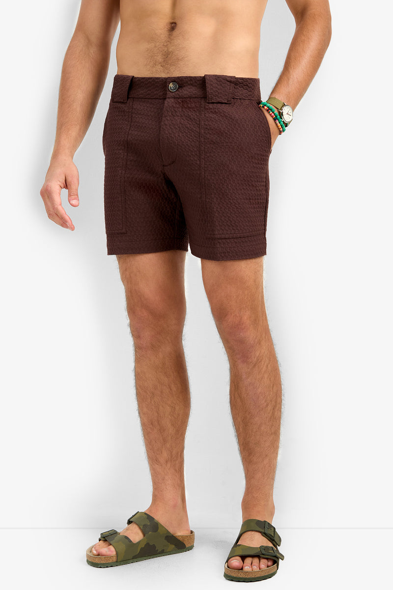 A man stands against a white background wearing the Randy Short in dark brown, green camouflage sandals, and a beaded bracelet. His left hand is in his pocket, and his upper body is not fully visible.