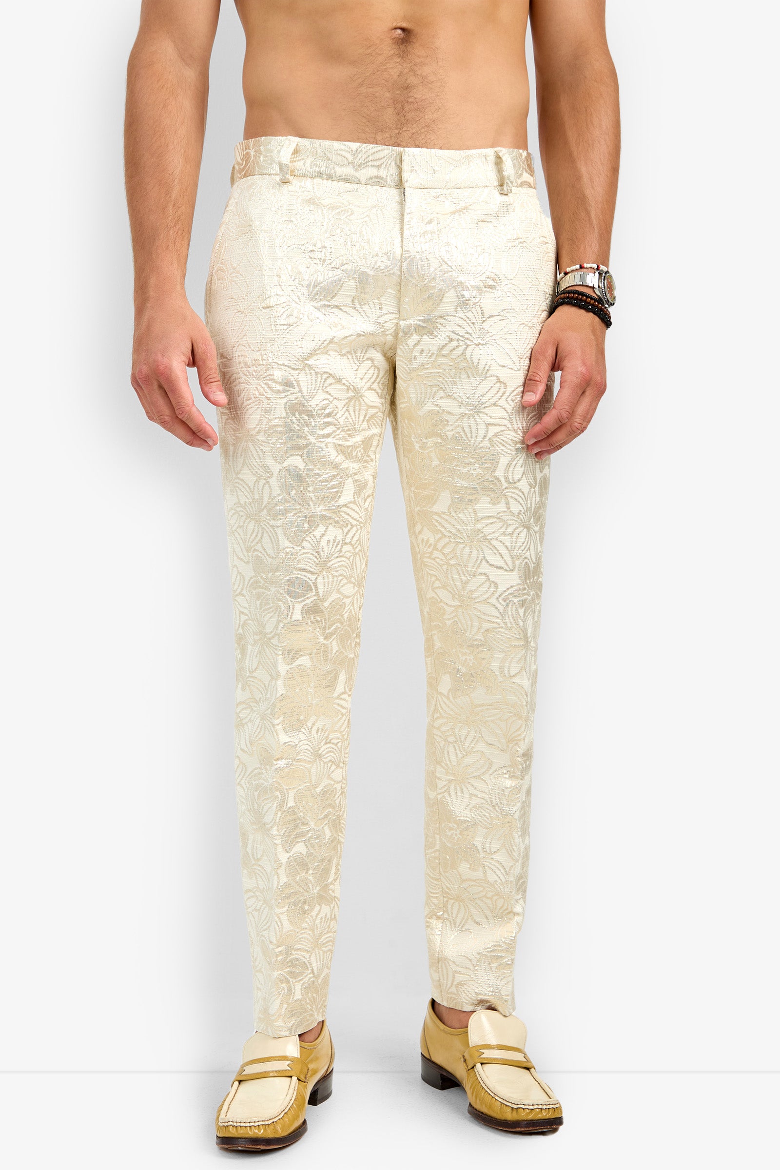 A shirtless man models the Clyde Slim Trouser—cream-colored, floral-patterned dress pants—with gold loafers against a white background. Only his body from shoulders to feet is visible, accessorized with a watch and bracelets on his left wrist.