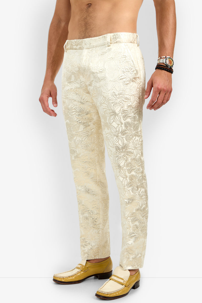 A shirtless man stands sideways in cream-colored Clyde Slim Trouser with a floral pattern and yellow loafers, accessorized with a watch and beaded bracelets.