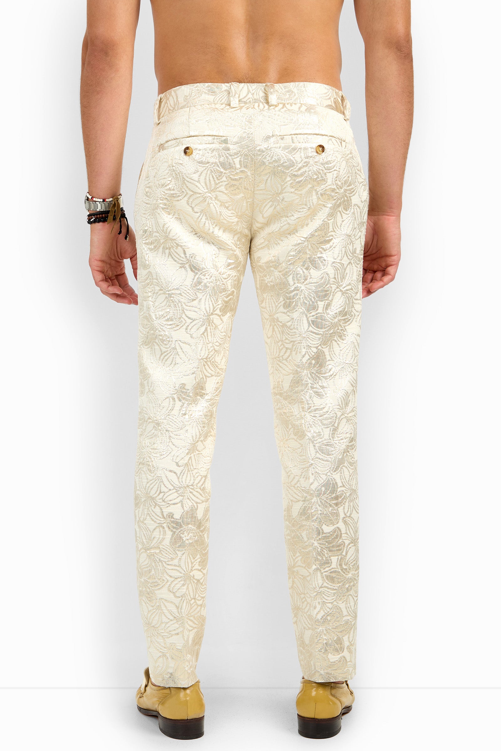 A shirtless man faces away, wearing the cream-colored Clyde Slim Trouser with a floral pattern and gold buttons, paired with beige loafers and bracelets on his left wrist. The background is plain white.