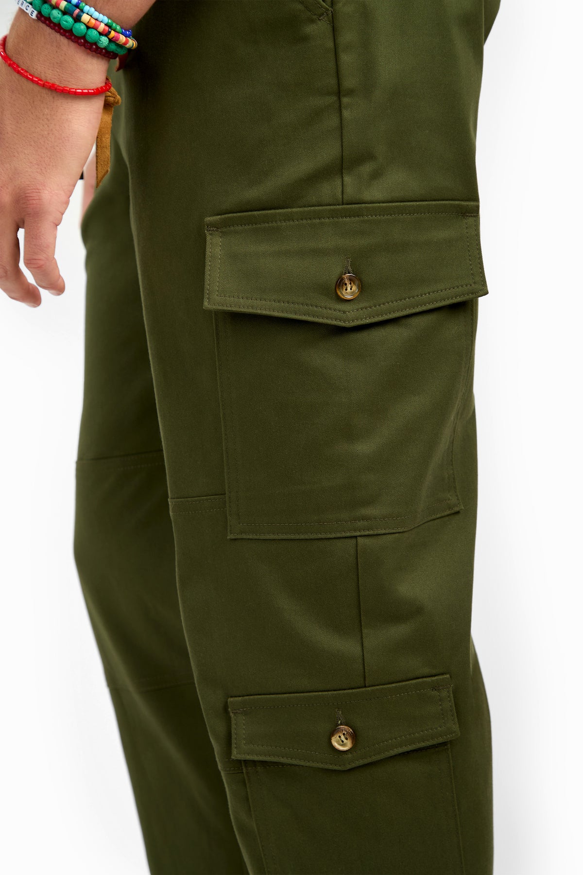 A close-up of a person wearing San Pedro Jogger olive green cargo pants with large buttoned pockets. Their left hand, adorned with colorful beaded bracelets, is visible at the side.