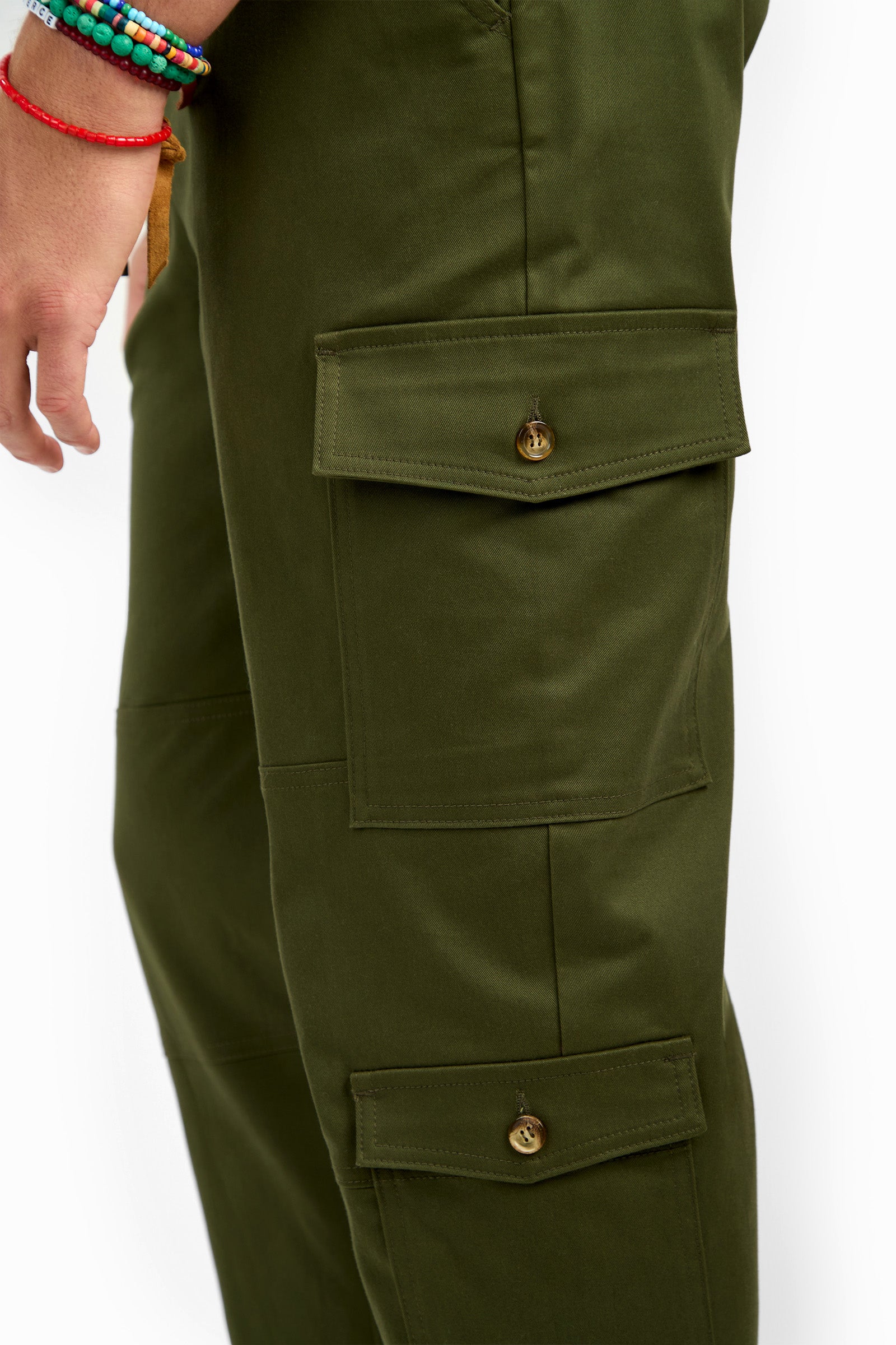 A close-up of a person wearing San Pedro Jogger olive green cargo pants with large buttoned pockets. Their left hand, adorned with colorful beaded bracelets, is visible at the side.