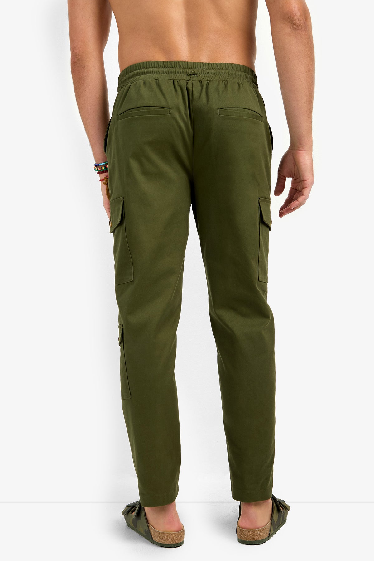 A person, shirtless and facing away, models the San Pedro Jogger—olive green cargo pants with back and side pockets—and brown sandals against a plain white background.