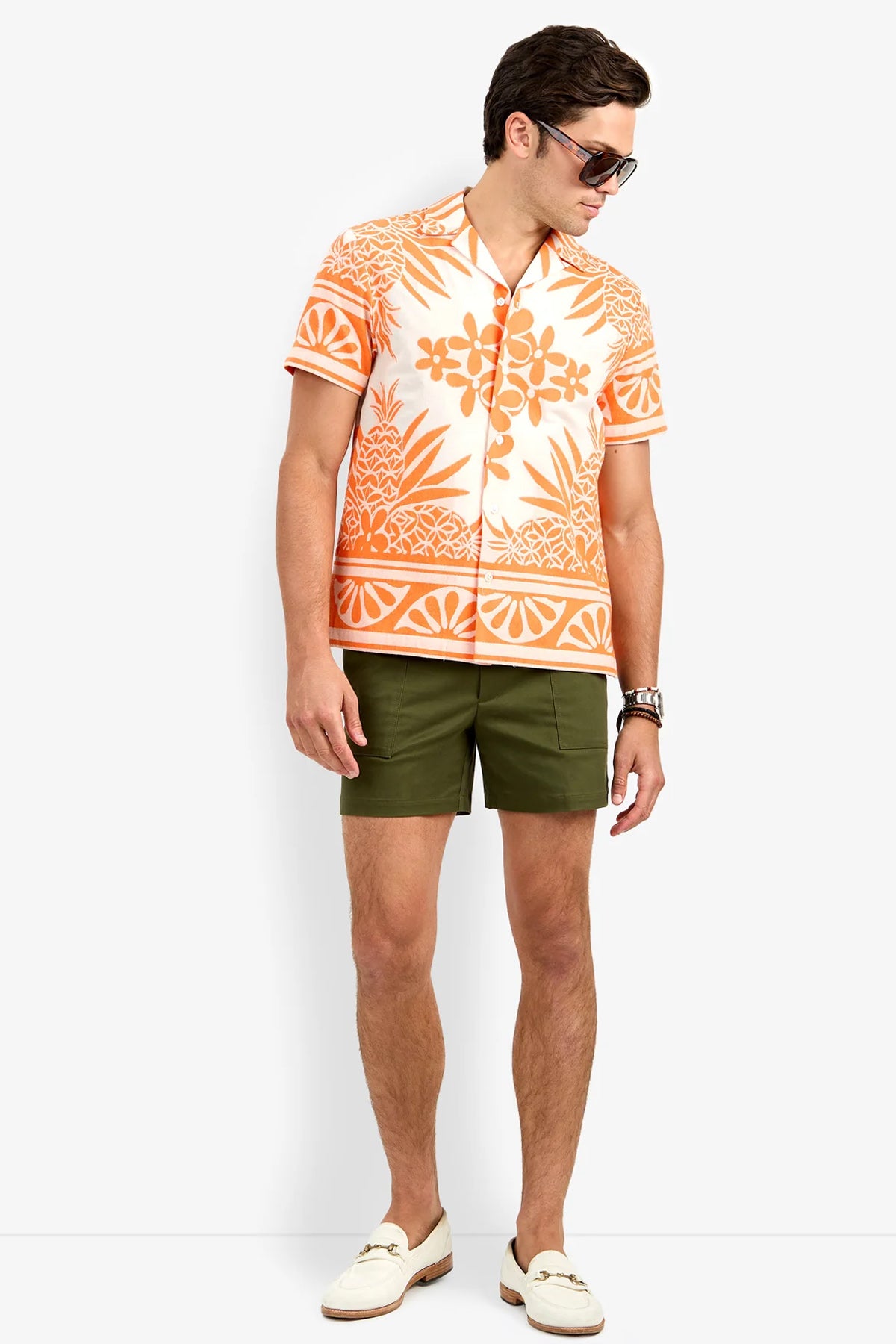 A man wearing sunglasses, an orange and white patterned short-sleeve shirt, the Will Rogers Short in olive green, and white loafers stands against a plain white background, looking down to his left.