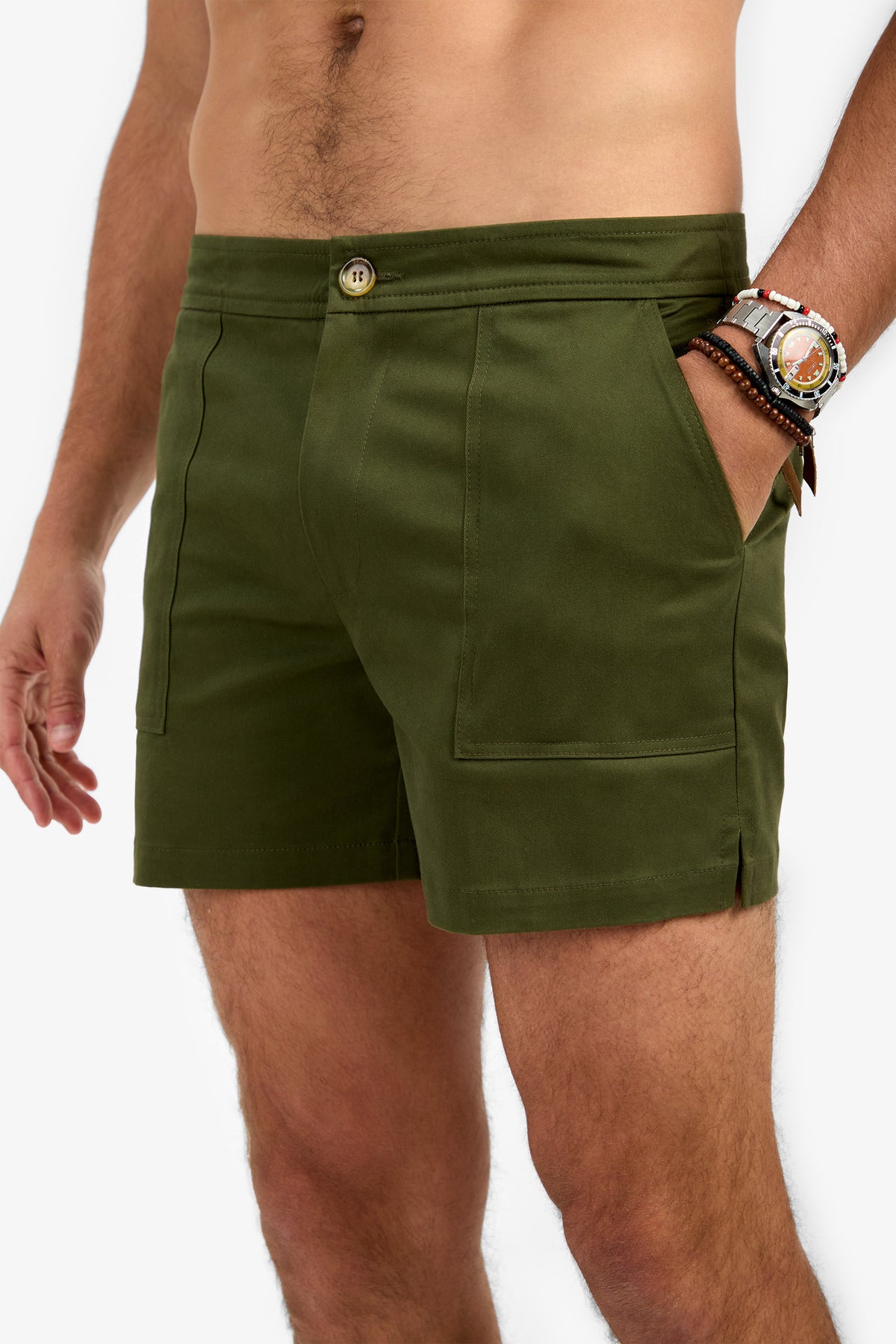 A man wearing the Will Rogers Short in olive green with front pockets stands against a plain white background, his left hand in a pocket. He sports a beaded bracelet and large watch on his left wrist; his upper body is not visible.