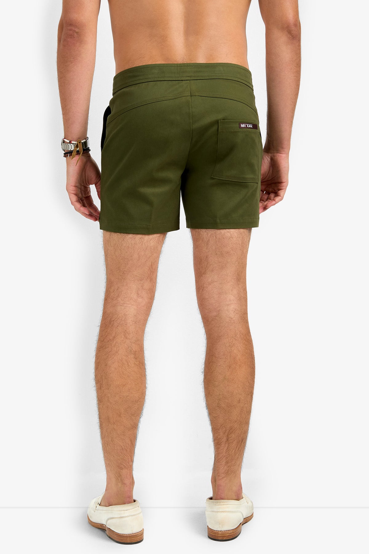 A man stands with his back to the camera, wearing Will Rogers Shorts in olive green, a wristwatch, and beige slip-on shoes. The shorts feature a right-side pocket. The background is plain white.