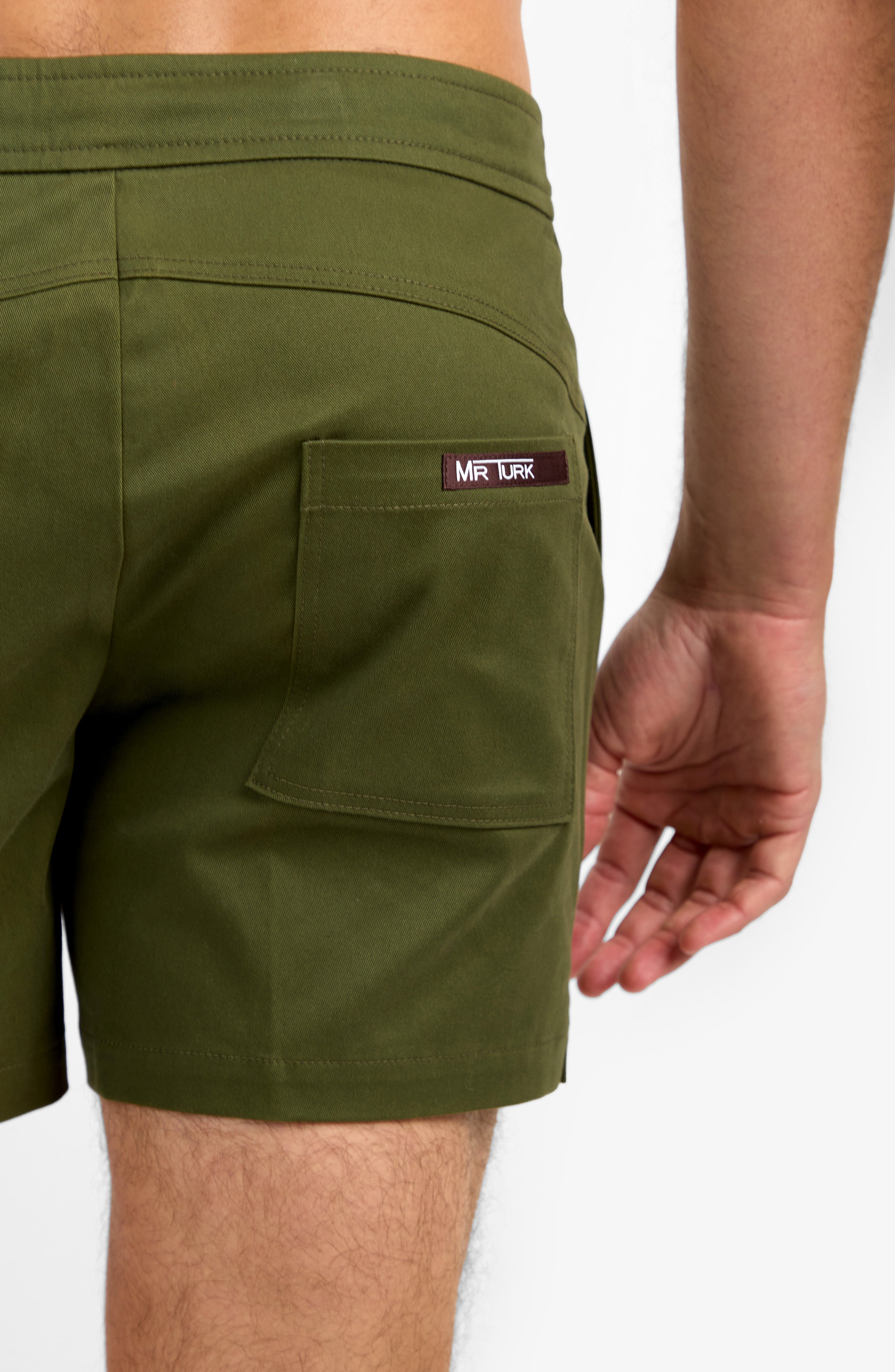 A person is shown from the waist down wearing Will Rogers Short in olive green, highlighting the back pocket with a MR TURK label and their right arm visible to the side.